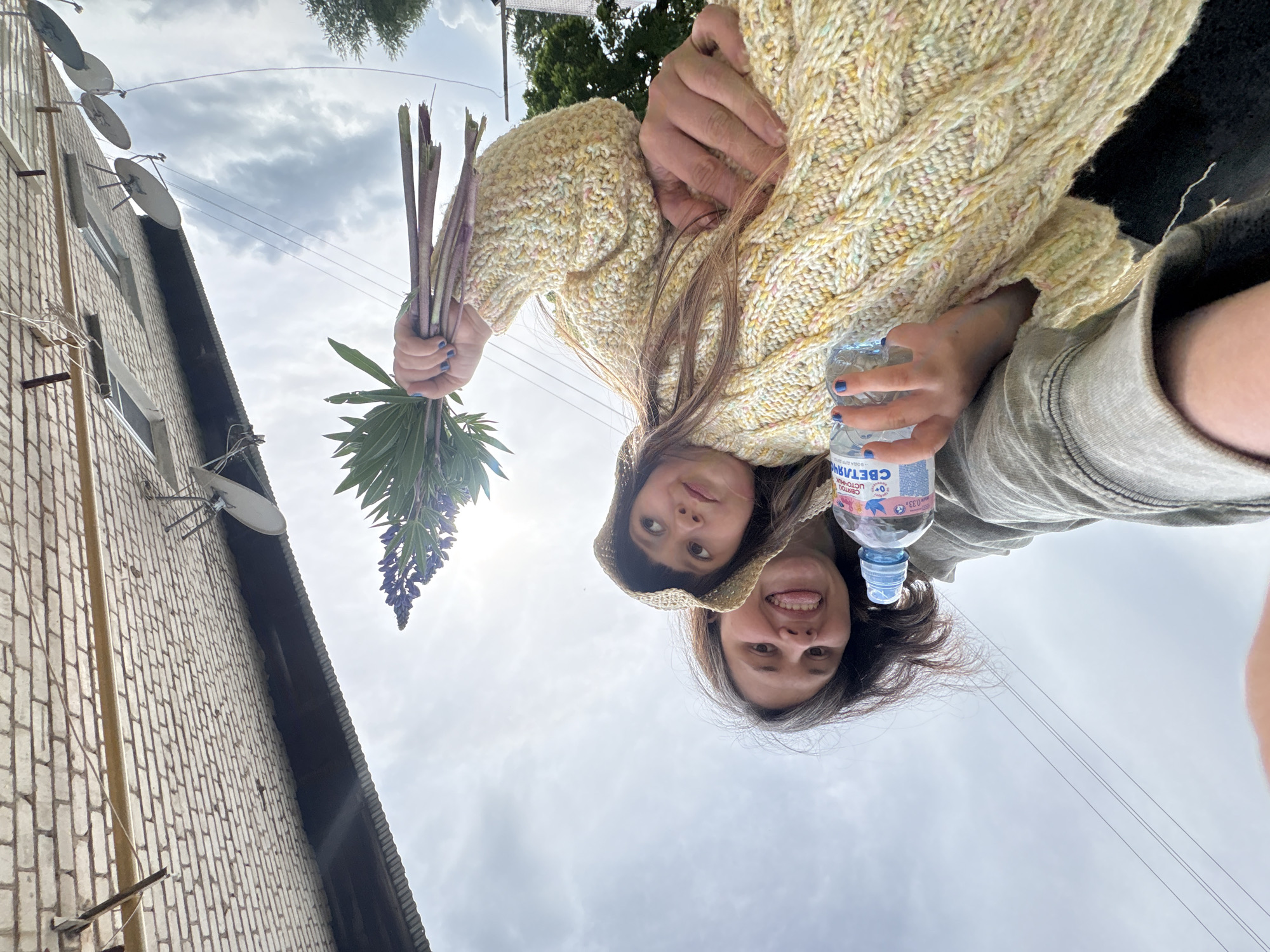 The image, taken from a low-angle perspective looking upwards, features two young women or girls set against a cloudy sky with parts of a brick building visible on the left. The scene is outdoors, likely in a residential area of Фоки, Russia, given the brick architecture, satellite dishes, and Cyrillic text on a bottle.
The woman on the left, closer to the viewer, wears a light yellow, cable-knit sweater and holds a bouquet of long-stemmed plants with green leaves and purple flowers. Her hair is long and brown. She has a gentle smile and looks towards the camera. The woman on the right, partially obscured behind the first, wears a gray top or hoodie and holds a clear plastic water bottle. She is smiling broadly and playfully sticking out her tongue. Both appear happy and relaxed.
On the left side of the frame, a light-colored brick building rises upwards, adorned with multiple white satellite dishes attached to its facade. Overhead, faint electrical wires traverse the sky, and glimpses of green tree foliage are visible at the top. The sky is predominantly cloudy, suggesting an overcast or partly cloudy daytime.
Visible text on the water bottle held by the woman on the right includes "СВЕТЛАЯ" (Svetlaya), "ИСТОЧНИК" (Istochnik), and "0,33 л" (0.33 liters), along with other smaller, less discernible Cyrillic words. The overall impression is a casual, cheerful moment captured outdoors.