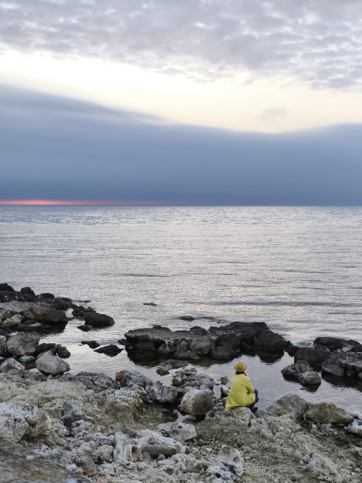 A wide shot captures a person wearing a yellow rain jacket and hat, sitting on a rocky shore, gazing out at the sea. The sky above is overcast, with a band of pink visible on the horizon line where the sky meets the water. The sea is calm with a slight shimmer on the surface, reflecting the gray sky. The rocky shore in the foreground consists of large, light-colored rocks interspersed with dark, wet stones closer to the water's edge. The scene is a quiet, contemplative moment at what could be a seaside location. The location is Sevastopol, Ukraine.