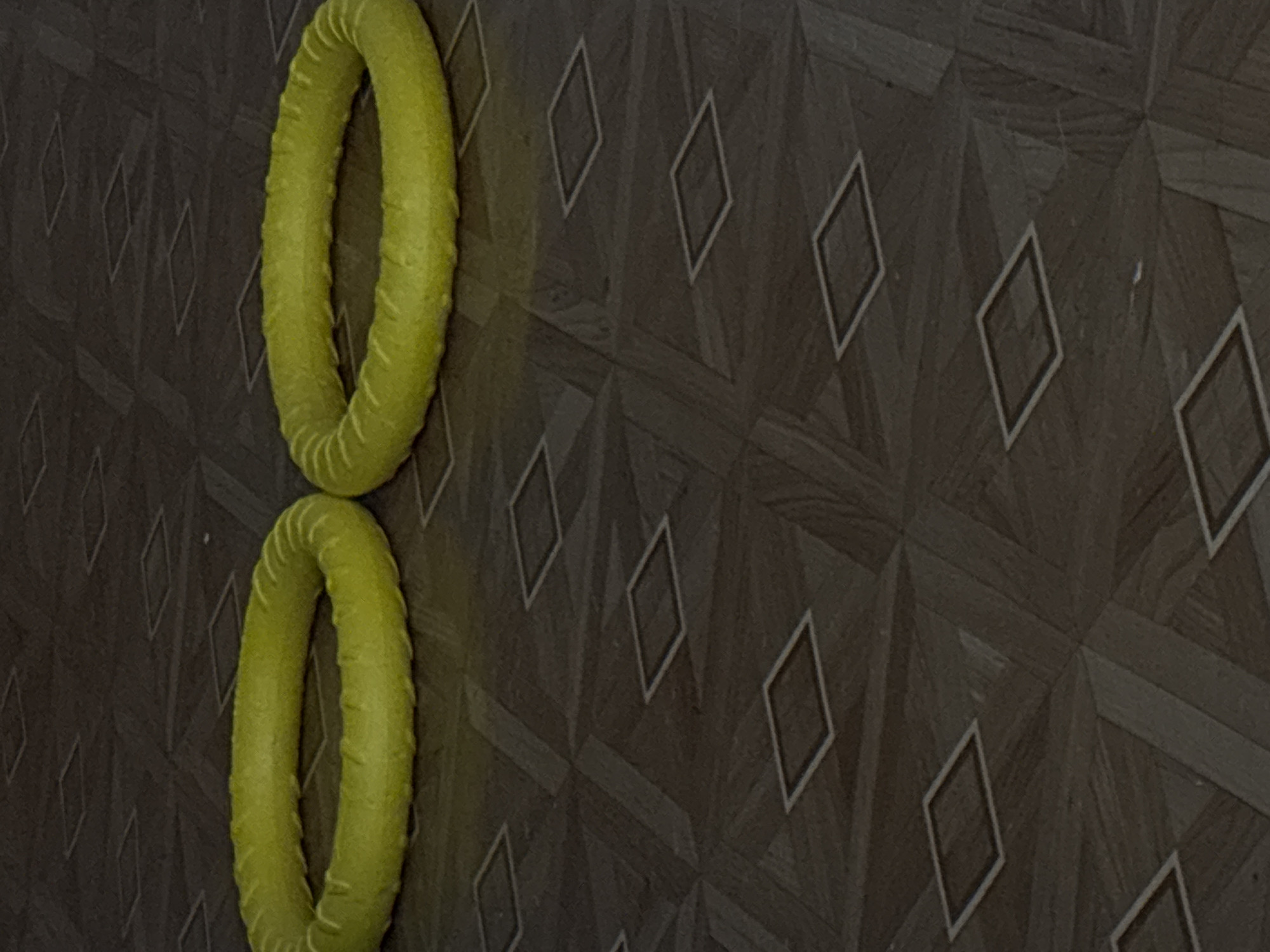 Two bright yellow, round, textured dog chew toys are positioned on a dark brown, patterned floor. The floor has a parquet design with diamond shapes outlined in white. The toys are side-by-side, with the left toy slightly closer to the viewer and taking up more of the frame. The lighting is dim, creating shadows that highlight the texture of the toys and the floor. This scene is not specific to any city or location within Saint Petersburg, Russia, as the environment depicted is generic and could be found in many indoor settings. There are no people, animals, or specific identifying landmarks present.