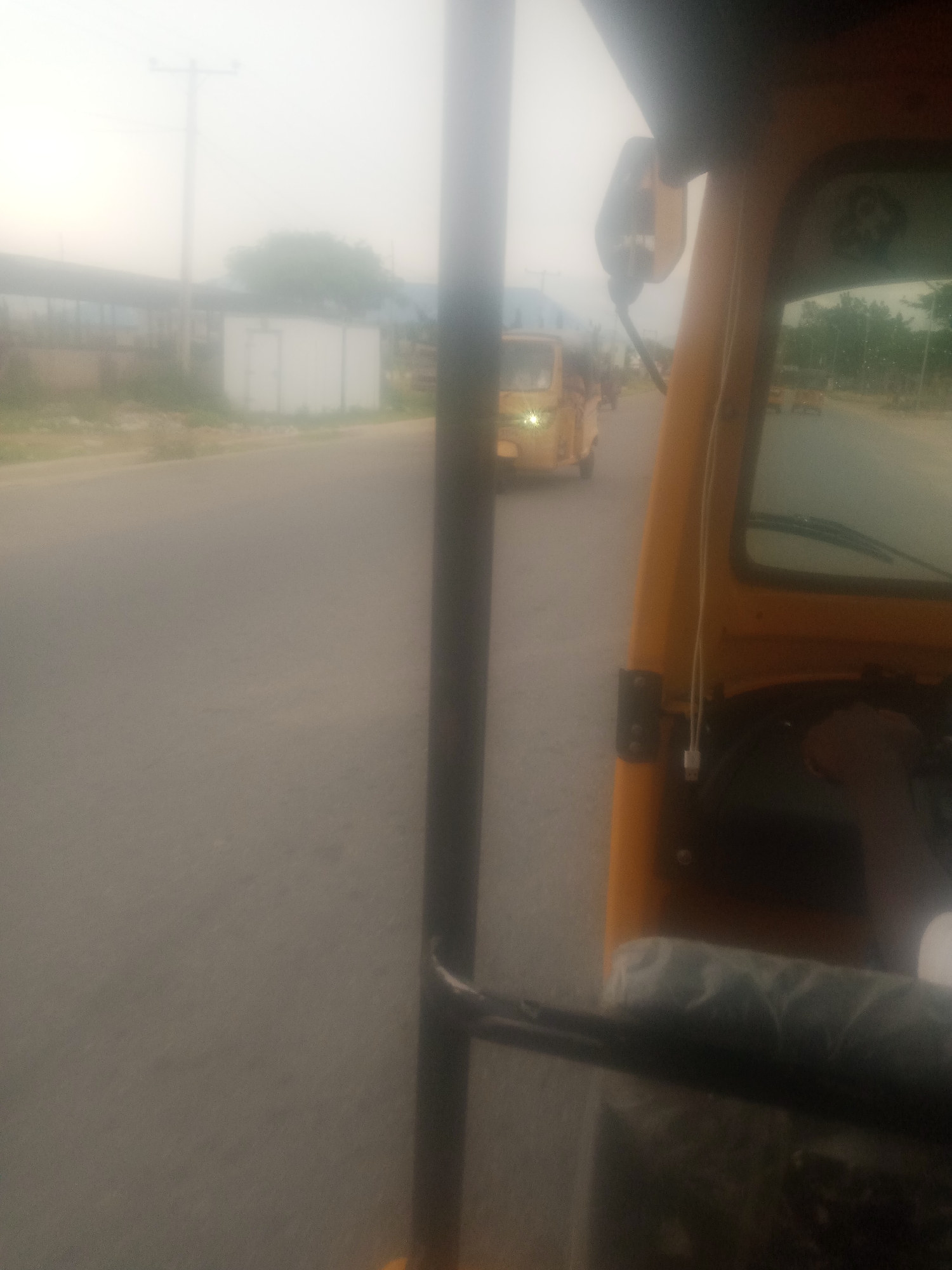 A slightly hazy, wide shot captures a view from within a yellow auto-rickshaw as it travels down a road in Jalingo, Nigeria. The auto-rickshaw is positioned on the right side of the frame, with its orange exterior and interior visible. The driver's side mirror is mounted on the windshield. In the distance, other auto-rickshaws are seen on the road. To the left of the road, a white structure, possibly a building or container, is visible, along with a utility pole. The sky is overcast, contributing to the hazy appearance of the scene. The road is paved, and the surrounding environment appears to be an urban or semi-urban area.