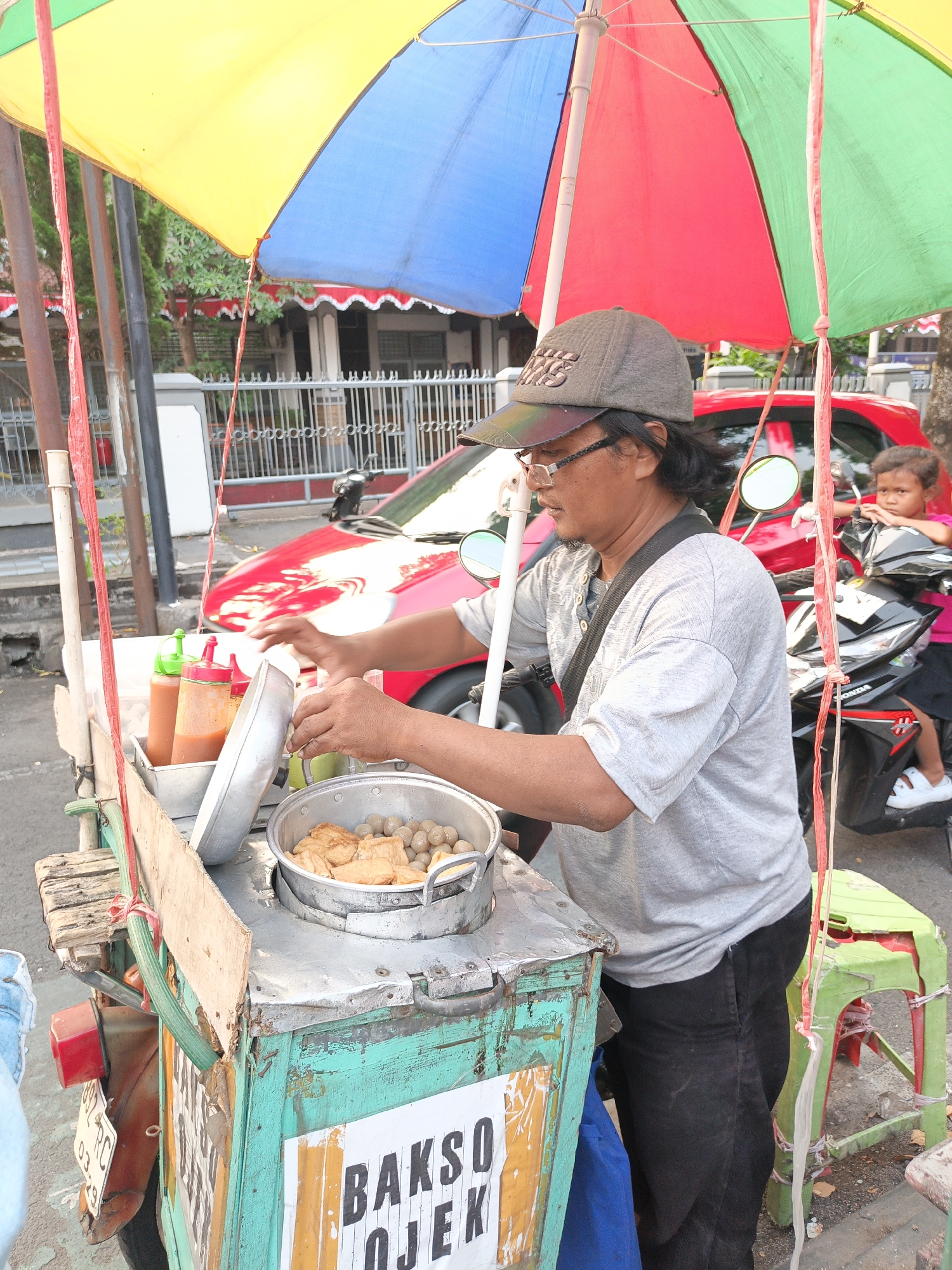 An Indonesian street vendor in Klaten, Indonesia, is preparing food at a cart. The vendor, wearing a grey t-shirt and a grey baseball cap, is seen with their hands over a metal pot containing meatballs and fried tofu. The cart is adorned with a colorful umbrella, yellow, blue, and red panels. Two condiment bottles, one red and one orange, are placed next to the pot. In the background, a red car and a black motorcycle are parked. A young girl, wearing a pink shirt and a black skirt, sits on the motorcycle, observing the scene. The cart itself is made of wood and metal, with a turquoise and green painted base. A sign on the cart reads "BAKSO OJEK." The scene appears to be set on a street, with a building visible in the distance.