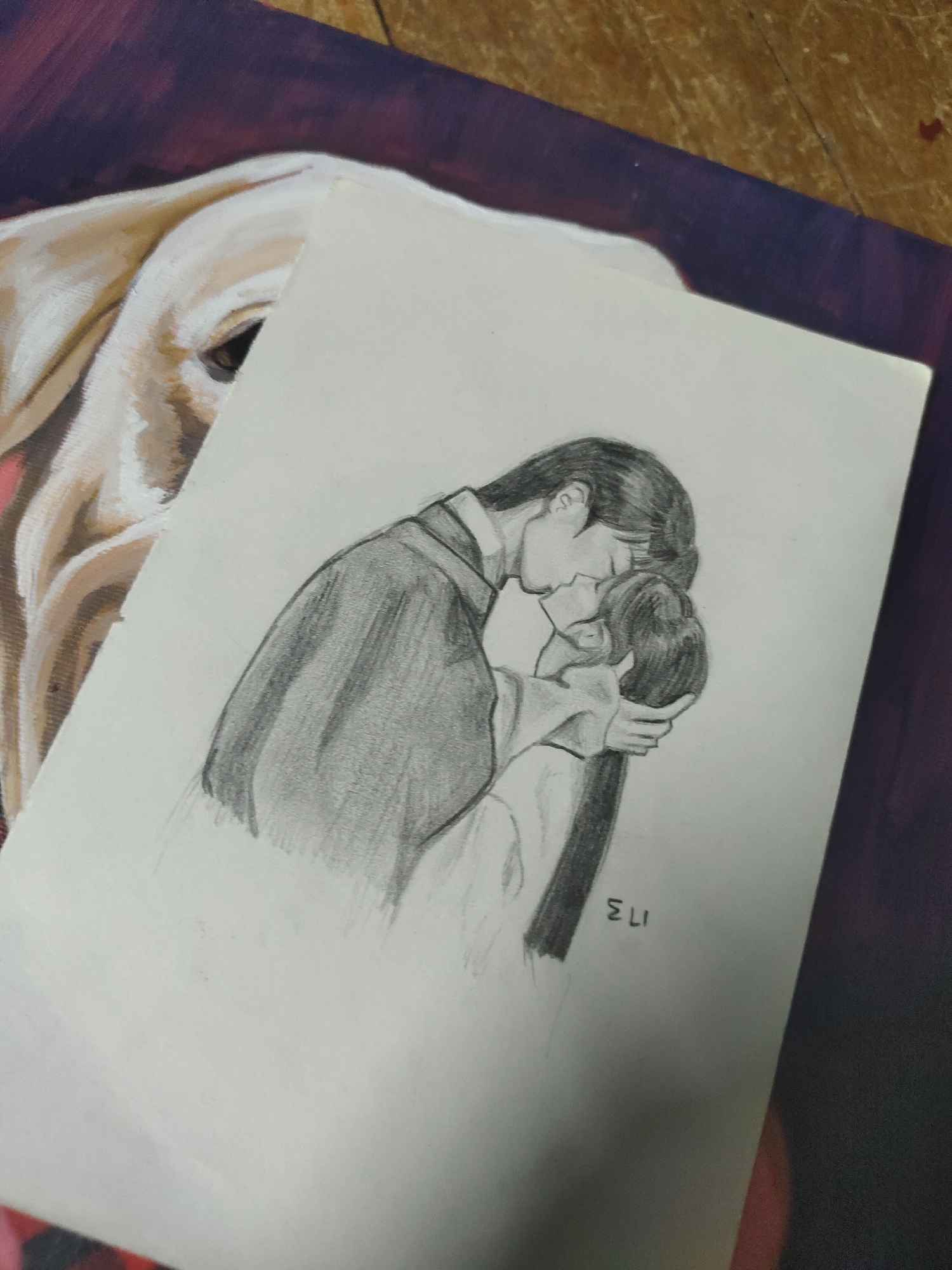 The image displays a white sheet of paper featuring a monochrome pencil drawing, diagonally positioned on a larger surface. The drawing depicts two figures, a man and a woman, embracing and in profile, kissing. The man, on the left, has short dark hair and is wearing what appears to be traditional or stylized clothing with broad sleeves. The woman, on the right, has long dark hair and is wearing similar attire. Her hand is visible on his face or neck. The signature 'ELI' is written in the lower right portion of the drawing.
Beneath the drawing, a portion of a colorful painting is visible. On the left, the eye and part of the head of a light-colored animal, resembling a dog (possibly a Labrador), are depicted. The animal's fur is rendered in shades of cream and beige. To the right and above the paper, a dark reddish-purple background or fabric is visible, along with a narrow strip of brown, textured surface, indicative of wood, at the top right corner. The lighting suggests an indoor setting.
