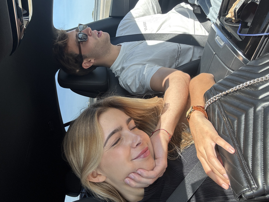 An interior view of a vehicle shows two individuals, a female and a male, in the front seats during daytime. The location is Santarém, Portugal.
The female is seated in the passenger seat on the left, secured by a black seatbelt. She has blonde hair and is depicted with her eyes closed, a soft smile on her face, and her right hand supporting her head. Her left hand rests on a black quilted handbag with a silver chain strap. She wears a dark, ribbed top, a thin red string bracelet on her right wrist, and a gold-toned rectangular watch with a brown leather strap on her left wrist.
The male, in the driver's seat on the right, is also secured by a black seatbelt. He has brown, curly hair and wears dark sunglasses with brown frames. His mouth is slightly open, and he appears to be looking forward, operating the vehicle. He wears a light-colored short-sleeved t-shirt with dark text, which includes phrases like "THE HIDDEN SYMPHONY" and "EXPLORING THE ETHNOBOTANICAL PRINCIPLES IN ART, AND DESIGN."
The car's interior features black upholstery and a dark headliner. A center console is partially visible, showing a blue charging cable connected to an unseen device. Through the windows, a clear blue sky with some distant white clouds and what appears to be a multi-lane road or highway are visible, indicating the vehicle is in motion. Sunlight illuminates the interior.