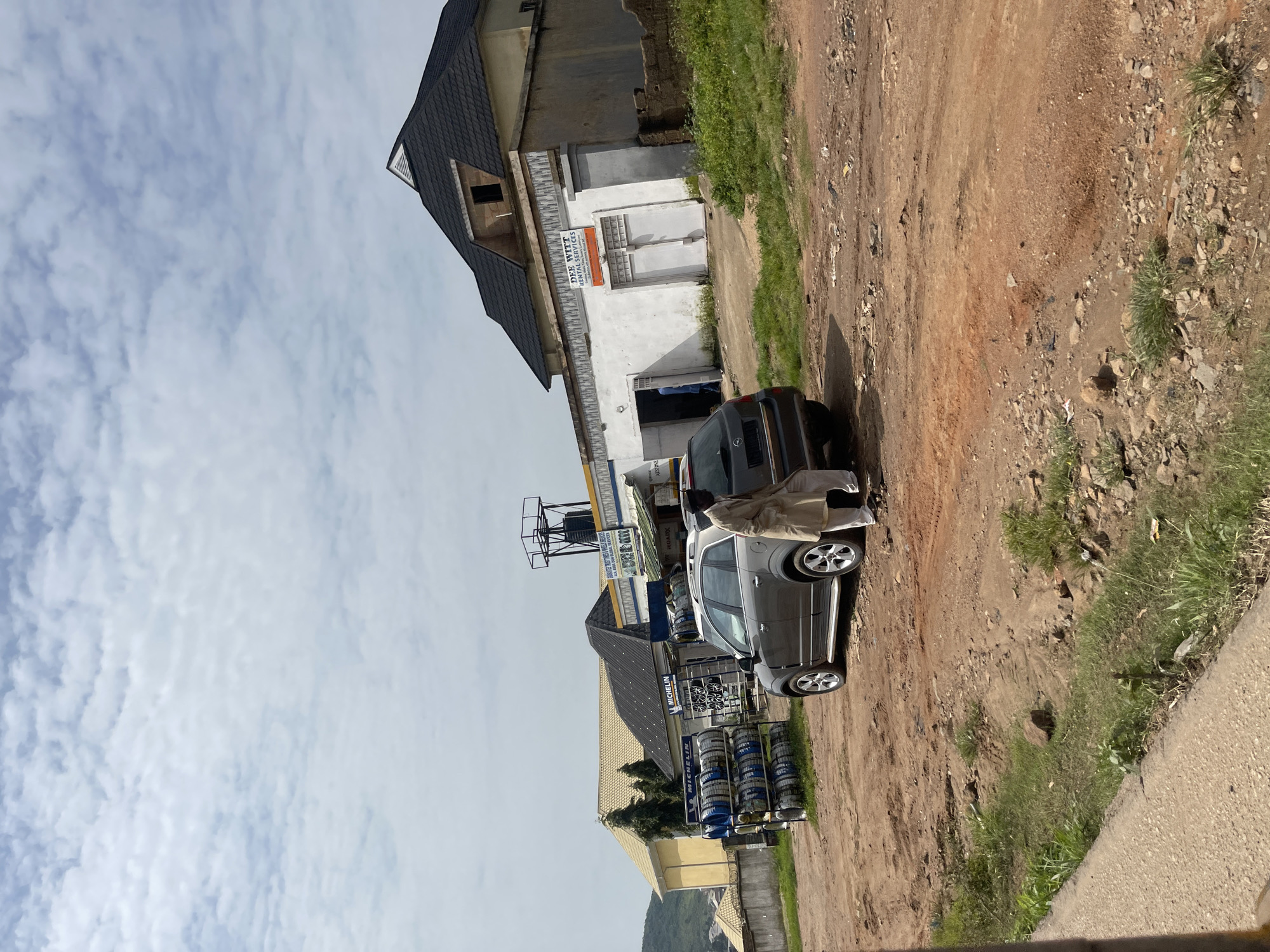 This image depicts an outdoor scene in Gwong, Nigeria, featuring a street with a dirt and gravel road. In the foreground, a gray SUV is parked, with a man dressed in a tan robe and white trousers standing beside its rear. To the left of the SUV, a rack displays several tires, some with blue and white branding that appears to read "MICHELIN."
Behind the SUV and tire rack, a building with a white facade and a dark, sloping roof is visible. A sign on this building reads "DEE WITT RENTAL SERVICES." Further in the background, another building with a yellowish facade and a pitched roof can be seen, along with a water tower. The sky is partly cloudy. The overall environment suggests a commercial area possibly related to automotive services.