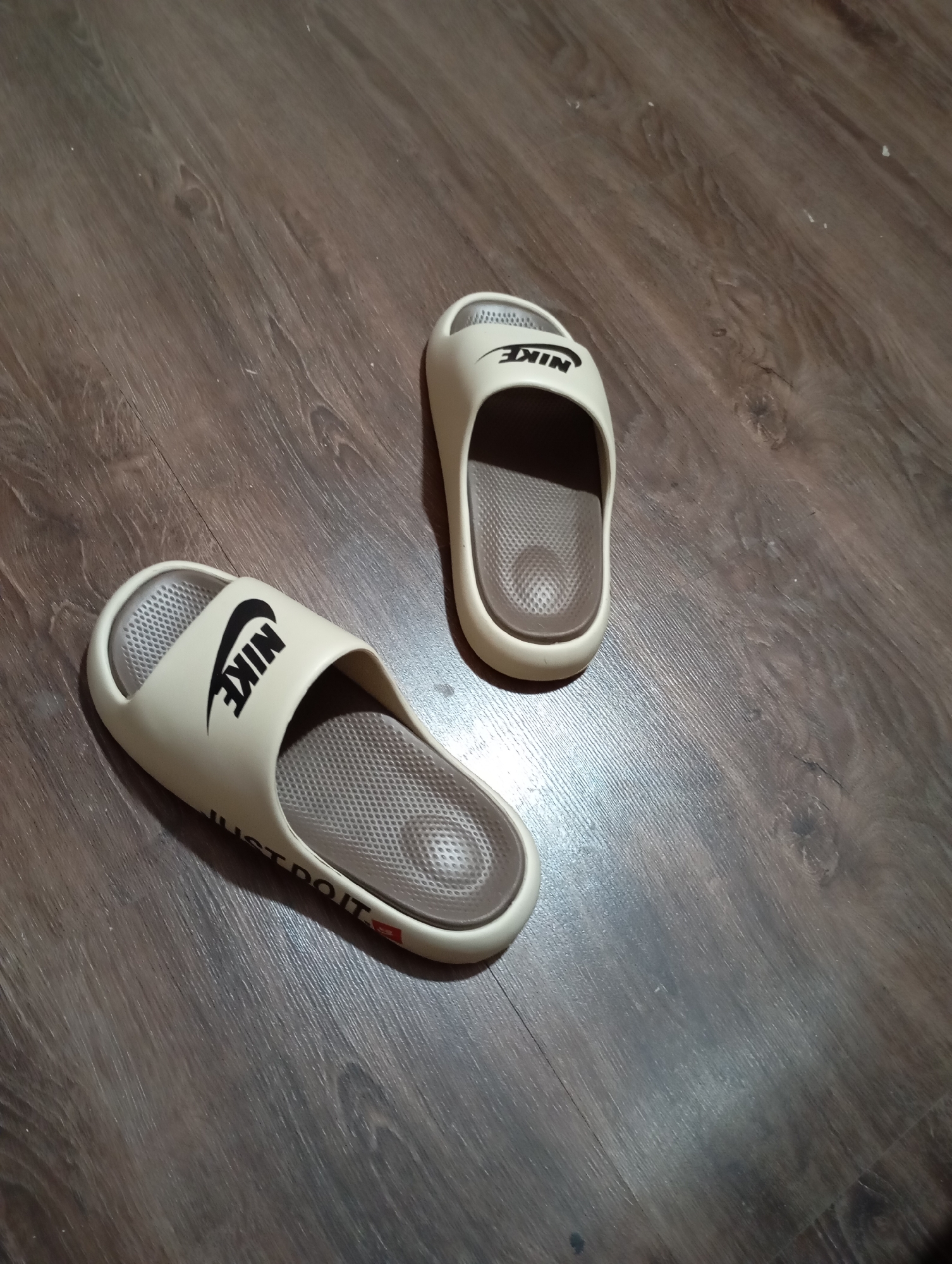 Two beige Nike slides are resting on a dark wood-grain floor. The slide on the left is closer to the viewer and displays the Nike logo in black on the strap, and the text "JUST DO IT" along the side. The slide on the right is further away and also shows the Nike logo on its strap. The floor appears to be an indoor surface, possibly a living space or entryway. There is no indication of the specific location within Almaty, Kazakhstan, as the image does not contain any identifiable landmarks or context clues.