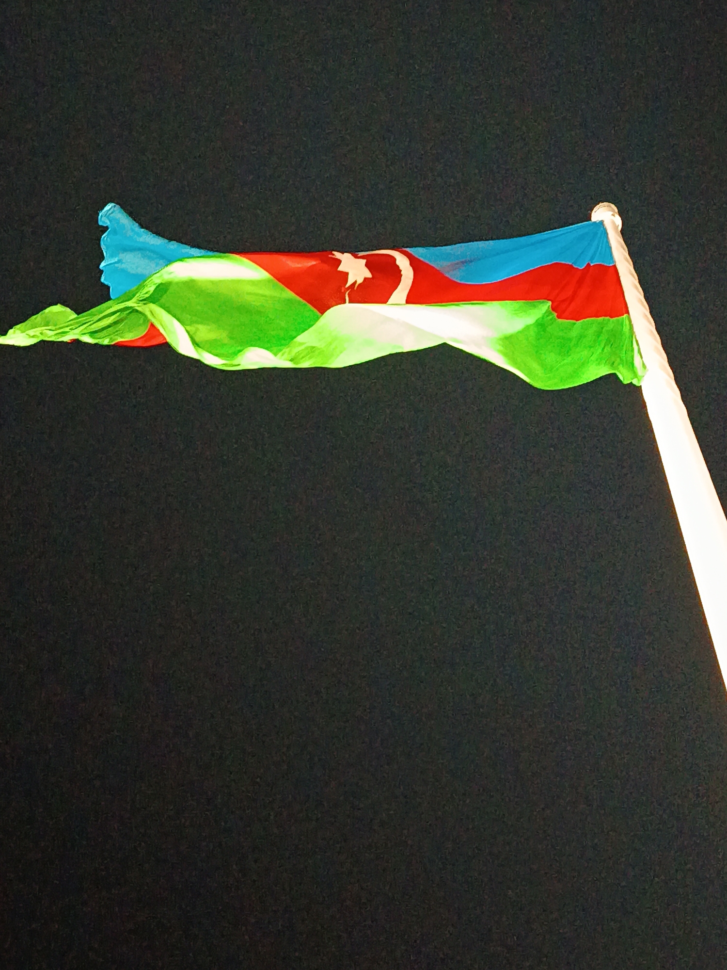 An Azerbaijani flag is flying against a dark sky. The flag is composed of three horizontal stripes of blue, red, and green, with a white crescent moon and star in the center of the red stripe. The flag is partially unfurled and appears to be in motion, with some rippling and folds visible. A portion of a white flagpole is visible on the right side of the image. The scene is likely outdoors, given the presence of a flag and the dark, presumably nighttime, sky. No people or specific landmarks are visible. The location is identified as Sabail Raion, Azerbaijan.