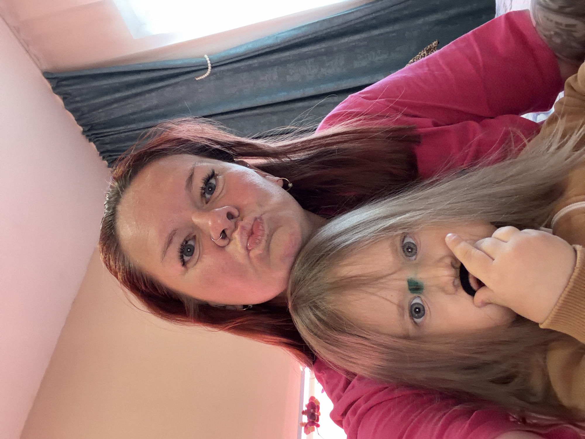 A selfie-style image shows two individuals. An adult with auburn hair, a nose ring, and wearing a pink top is making a kissing face. A child with blonde hair is holding a black elastic band up to their nose, with a green mark on their forehead. The background shows a window with a curtain, and a peach-colored wall. The location is undetermined; the city of Chaikovsky, Russia is not discernible from the image.