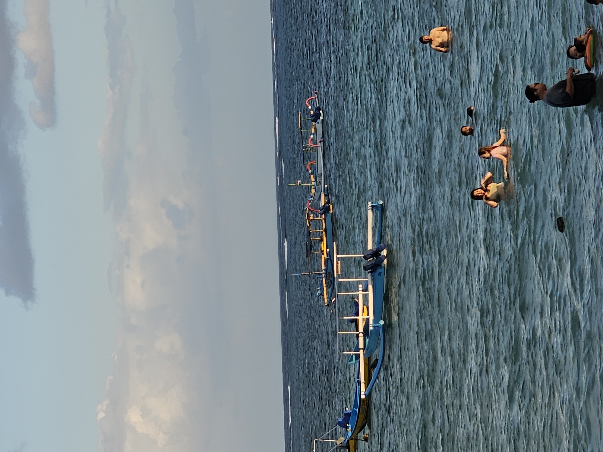 This image captures a scene at a beach in Denpasar, Indonesia, likely in the late afternoon or early evening given the lighting. The foreground features several people bathing and swimming in the ocean. Among them is a man standing chest-deep in the water, and to his right, a woman and a young girl are visible, with the girl appearing to be in a pink swimsuit. Further left, another woman is partially submerged. Closer to the foreground on the right, a man is standing in the water, holding a cup and facing away from the camera, with two children near him, one of whom is wearing a colorful float. The middle ground of the image is occupied by several traditional Balinese fishing boats, known as jukungs. These boats are mostly blue and yellow and are moored or drifting in the calm sea. The background shows the vast expanse of the ocean meeting the horizon under a sky with scattered clouds, some of which are tinged with the warm hues of sunset. The overall atmosphere is tranquil and suggests a leisurely day at the beach.