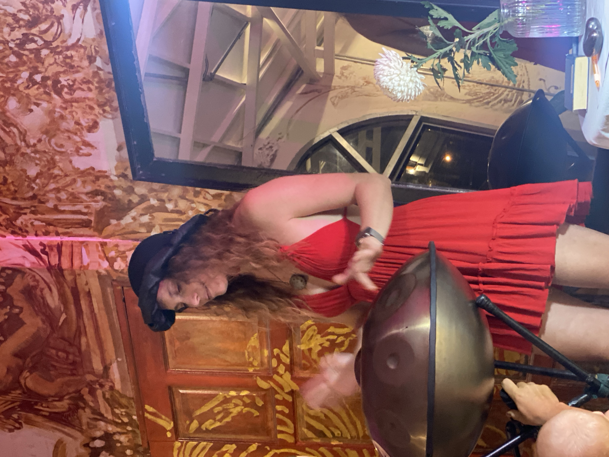 A woman wearing a red dress and a black hat is lying down, her head resting on a decorative surface. She has her eyes closed and a relaxed expression. Her right arm is extended, and her hand is positioned as if playing a musical instrument. In front of her, a large, spherical, metallic object with various indentations rests on a stand, suggesting it is a handpan drum. The background features an ornate wall with a mural-like design in earthy tones and gold accents. Above, a mirror reflects the ceiling structure, which appears to be a combination of beams and painted surfaces. A white flower with delicate petals is visible near the top of the frame, attached to a green stem. The lighting in the scene is somewhat dim, with hints of pink and blue light suggesting an indoor ambiance, possibly a performance or a creative space. The overall mood is serene and contemplative. There is no visible text.