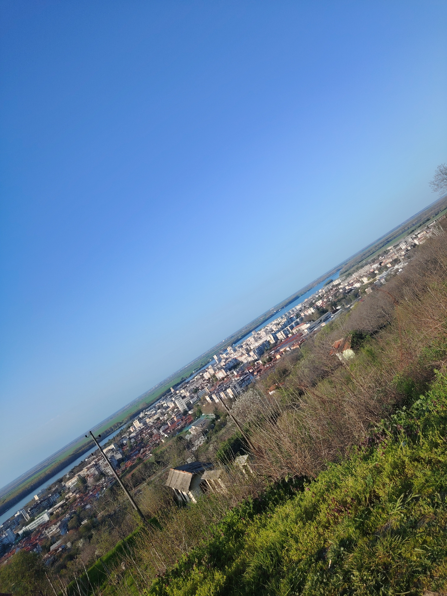 A wide-angle, slightly tilted, daytime shot shows a city with a river running alongside it. In the foreground, the ground slopes downwards, covered with low-lying green plants and dry bushes. Further down the slope, a small, dilapidated wooden structure with a rusty-colored roof is visible. An electric pole, with wires running through it, is also seen near the structure. Beyond the immediate foreground, a cityscape unfolds, marked by various buildings, mostly of moderate height. A wide river is bordering the city on one side. Beyond the river, the landscape extends into flat, green fields, eventually meeting the horizon. The sky above is a clear, bright blue.