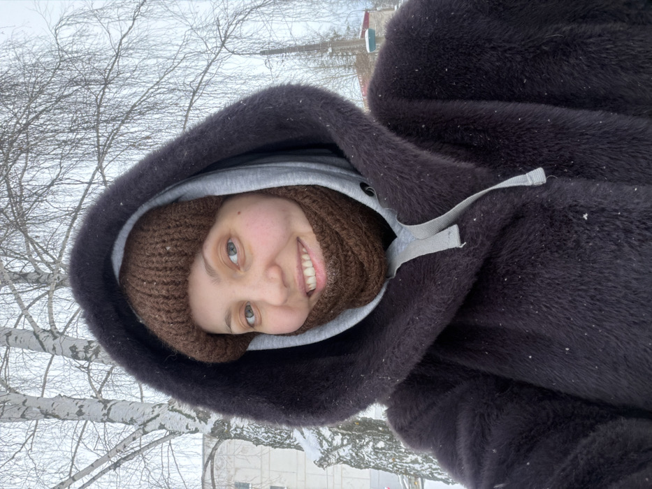 This image depicts a female individual in a snowy outdoor environment, consistent with winter conditions in Chaikovsky, Russia. The subject is centered, smiling directly at the camera. She is dressed in cold-weather attire, including a dark brown hooded coat with a visible grey hooded sweatshirt underneath, a dark brown knitted beanie, and a dark brown knitted neck warmer covering her lower face. Her eyes are light-colored. The background features bare deciduous trees, notably birch trees with white bark, with snow covering branches and the ground. A multi-story building is partially visible on the far left, and a tall industrial-style structure is present on the far right. Small white specks, consistent with falling snow, are visible on the subject's coat.