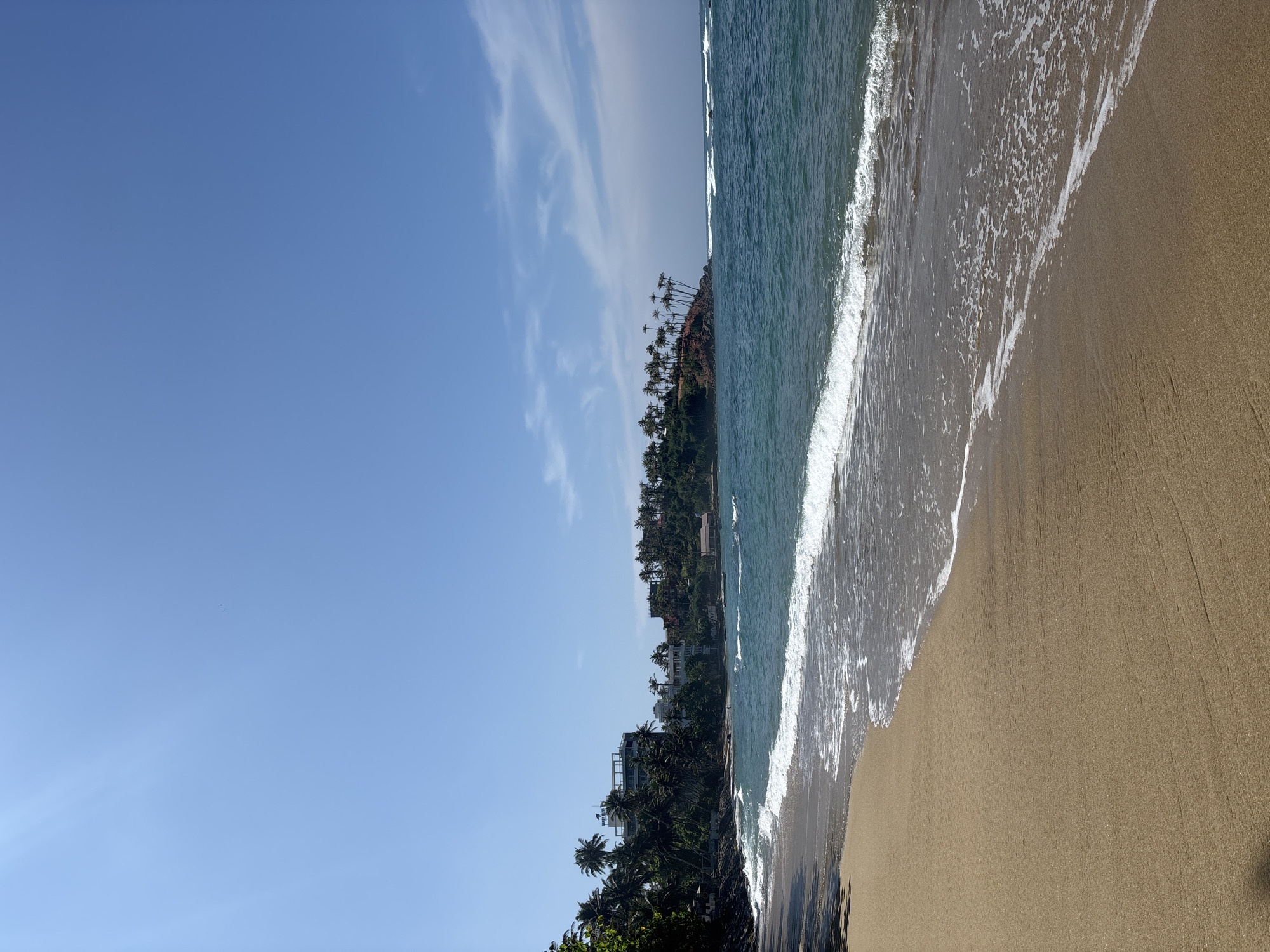 The image presents a beach scene in Thalaramba, Sri Lanka. A light tan sandy beach is on the right side of the frame with gentle waves from a teal-colored ocean washing onto the shore. On the left, a green coastline with palm trees and buildings ascends, with a clear blue sky overhead marked by wispy clouds. The scene appears serene and tropical.