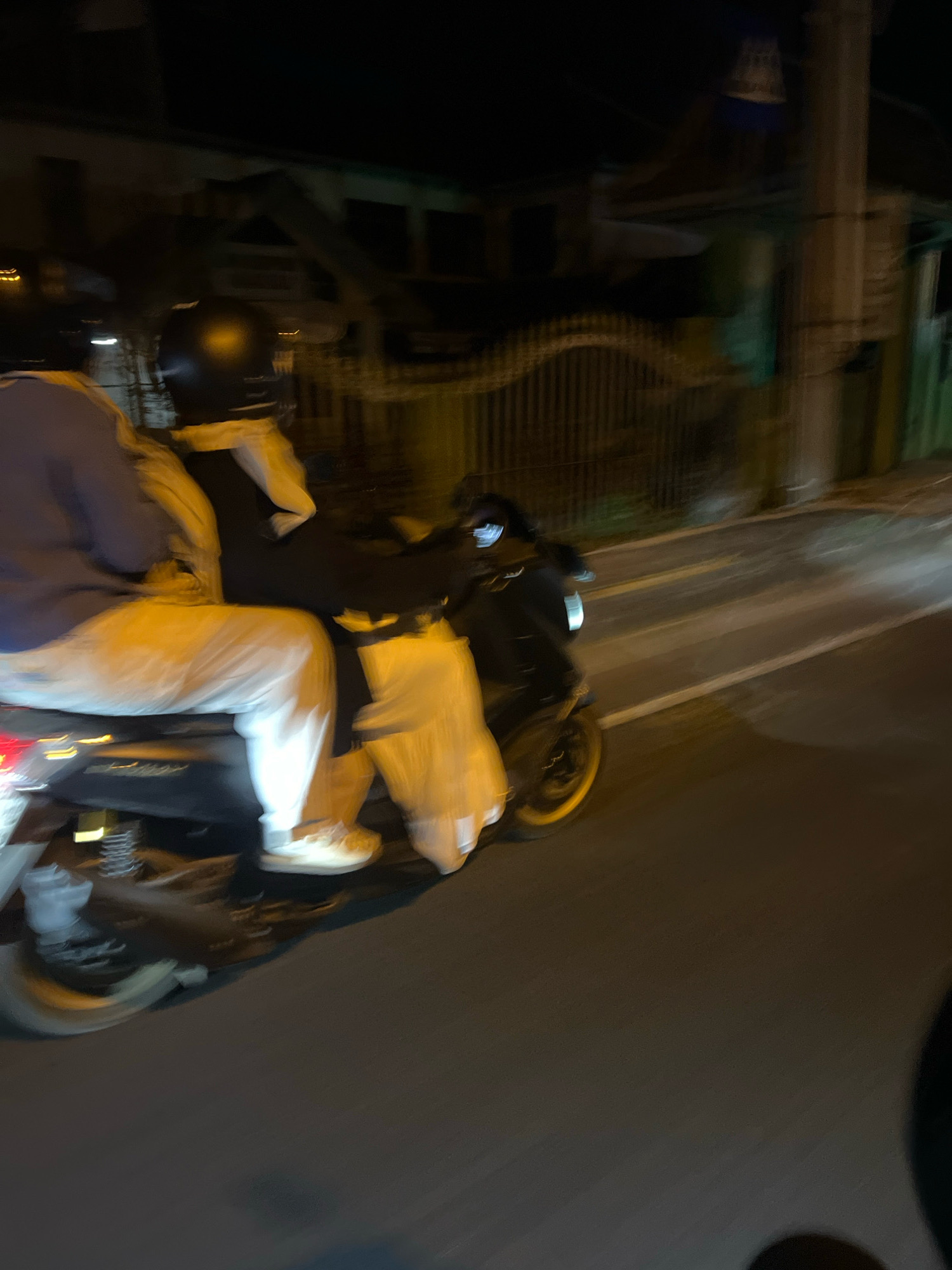 Two individuals are riding a scooter at night on a paved road in Magelang, Indonesia. The person in the front is wearing a dark helmet and a black jacket over a white shirt, while the person in the back is wearing a blue jacket and white pants. The scooter appears to be moving, as indicated by the motion blur. The environment shows buildings and a fence in the background, illuminated by streetlights and possibly lights from the buildings. The road has yellow and white lane markings.