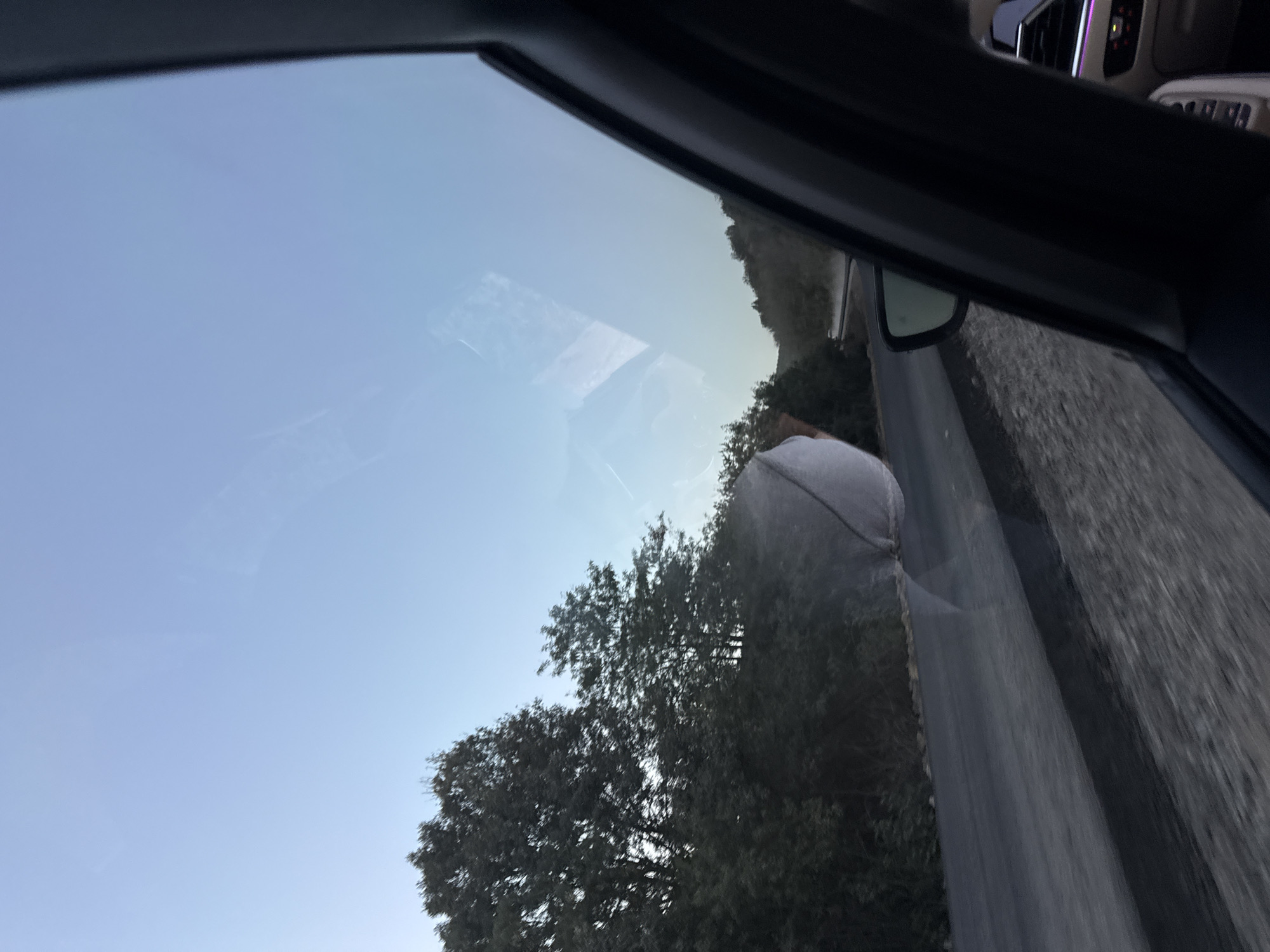 The image shows a view from inside a vehicle in Sărata-Mereșeni, Moldova. The perspective is from the passenger seat, looking out the side window. A road is visible, bordered by trees and foliage on the left side. The sky above is a light blue, with some reflections on the window. The interior of the car, including a side mirror and part of the dashboard with illuminated buttons, is also visible on the right. There is a slightly blurred, white, irregularly shaped object adhered to the exterior of the window. The overall scene suggests a travel scenario during daylight.