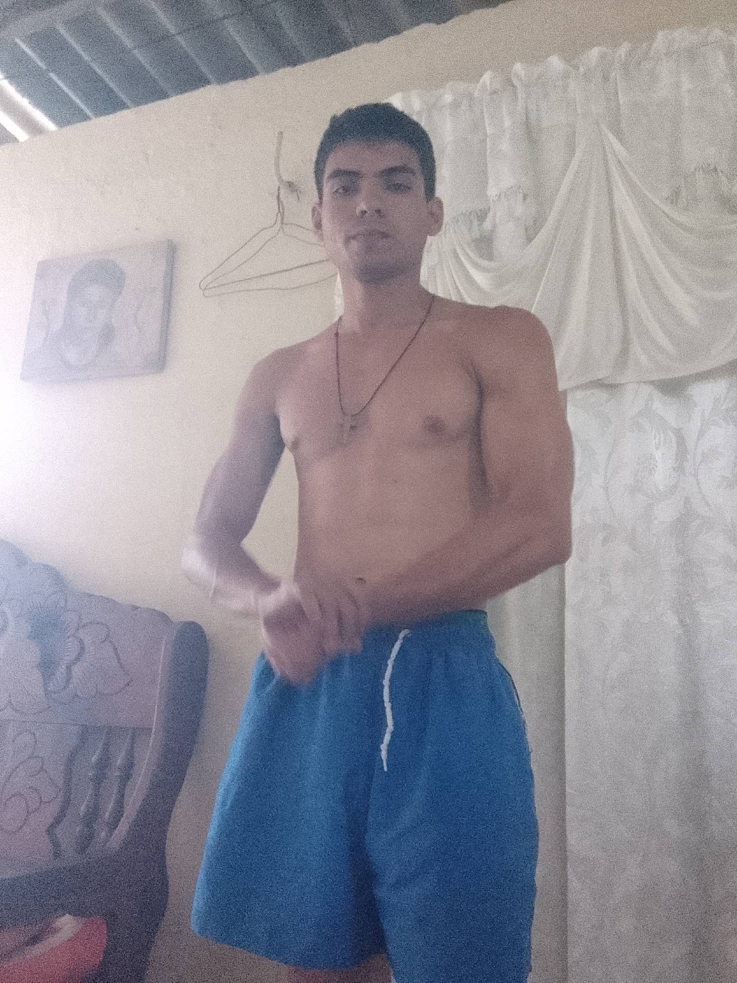 A shirtless man stands indoors wearing blue shorts with a white drawstring. He has dark hair, olive skin, and is wearing a necklace with a cross pendant. He is clenching his fists in front of his waist. Behind him, there's a white curtain, a framed picture on the wall, and a wooden clothes hanger. To his left, there is a carved wooden chair. The location is likely a private residence in Juan Griego, Venezuela, given the presence of typical Venezuelan furnishings and the lack of commercial or public features in the scene.