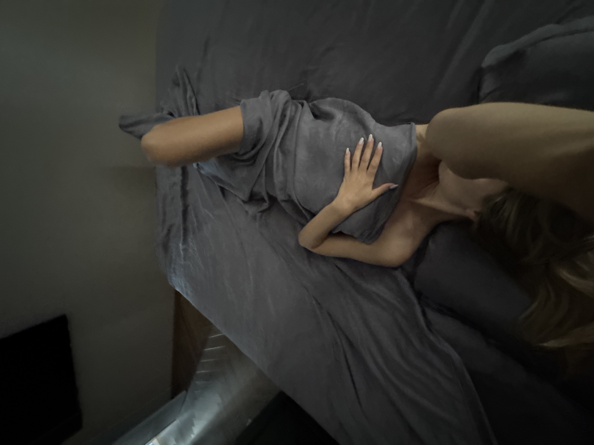 A woman is lying on a bed covered with gray sheets. She is wearing a gray towel wrapped around her. Her left leg is bent at the knee and extended upwards, while her right leg is bent at the knee and resting on the bed. Her left hand is resting on the towel, with her fingers extended. Her head is tilted back, and her eyes are closed. The room is dimly lit, with a window on the left side of the frame. The floor is made of hardwood.