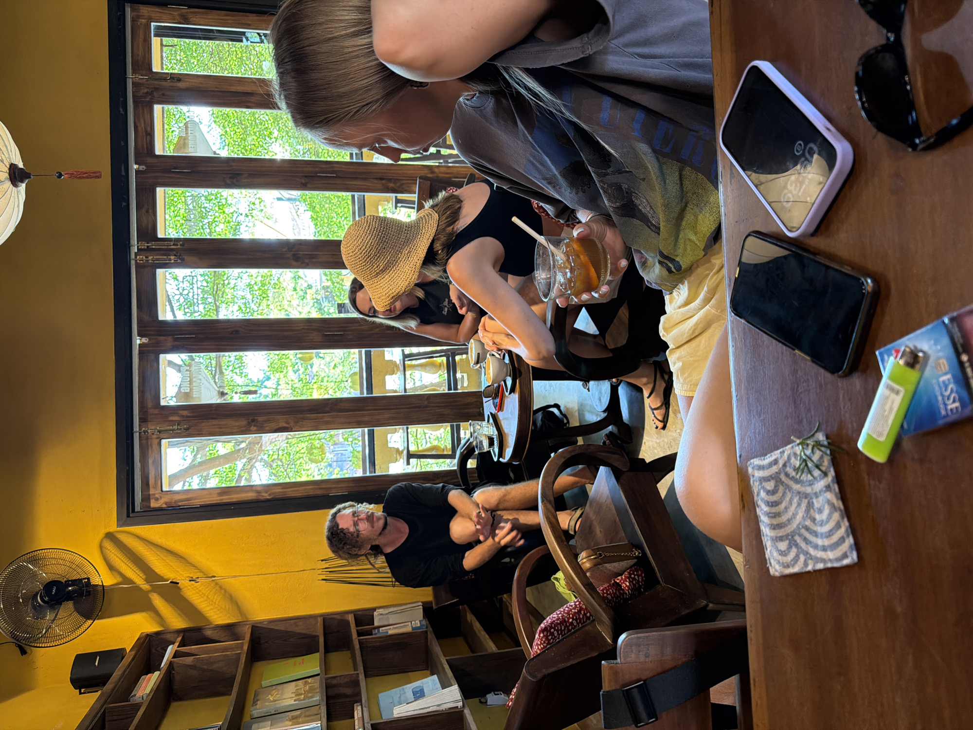 In a dimly lit cafe in Hội An, Vietnam, four individuals are engaged in conversation. The cafe's walls are a vibrant yellow, and large wooden-framed windows offer a view of lush greenery outside. A ceiling fan circulates the air above a group seated around a small table.
A man with curly hair and glasses is seated in a wooden armchair, his hands clasped in his lap. He wears a black t-shirt and shorts. Opposite him, two women are seated. One wears a straw hat and a black top, while the other, a blonde woman, is turned away from the camera, looking towards the man. She holds a glass of amber liquid with ice and a slice of orange. The fourth person, unseen, is implied by the presence of a phone and sunglasses on the table in the foreground.
The table also holds two smartphones, a pack of cigarettes, a green lighter, and a small patterned cloth with a sprig of rosemary. The cafe's decor includes wooden shelves filled with books and decorative items, and a traditional Vietnamese lantern hangs from the ceiling. The overall ambiance suggests a relaxed and casual gathering.