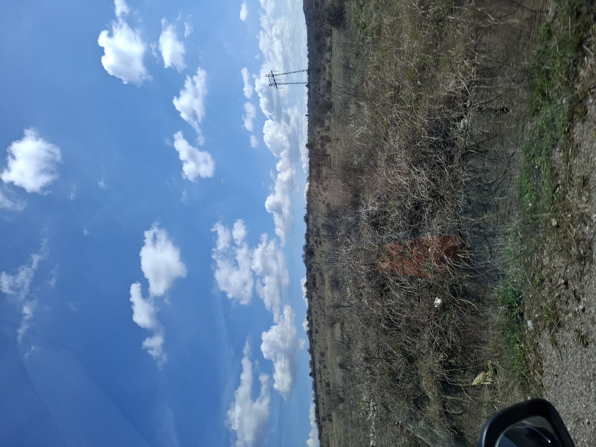 The image shows a rural roadside scene near Ponor, Bulgaria. The sky is bright blue with scattered white clouds. High-voltage power lines stretch across the landscape, supported by a pylon in the distance. The ground is covered in dry, scrubby vegetation with patches of green grass appearing along the edge of the road. A section of road is visible in the foreground. The photograph was likely taken from inside a vehicle as a side-view mirror is visible.