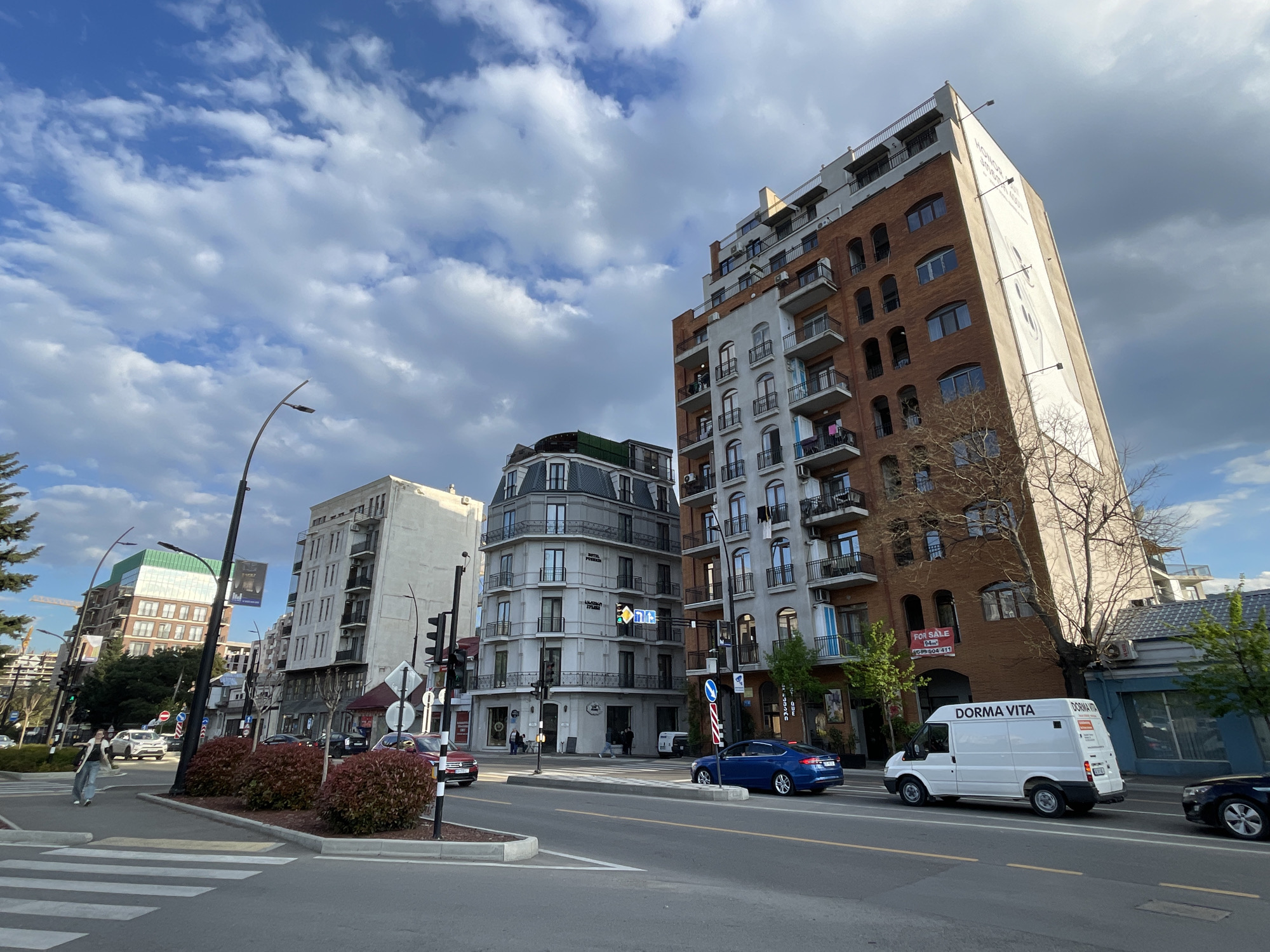 The image captures a dynamic street scene in Tbilisi, Georgia, under a bright, partly cloudy sky. The setting is an urban street lined with diverse architectural styles, featuring both modern and older buildings.
In the foreground, a multi-lane road is busy with vehicles. A white commercial van, prominently displaying "DORMA VITA" on its side, drives from right to left. Behind it, a blue sedan and other cars are visible. On the left side, a pedestrian is seen crossing a zebra crossing. The street is equipped with traffic lights, streetlights, and various road signs. A median strip with small red bushes separates parts of the road.
The buildings vary significantly. On the right, a tall, multi-story building with a mix of red brick and light-colored sections features numerous balconies. A large, vertical advertisement adorns its right side, displaying "HONOR" in English along with text in Georgian script, likely advertising a phone such as "HONOR MAGIC". A "FOR SALE" sign is also visible on this building, showing "94m²" and a phone number "599 604 411". In the center, a distinctive grey, ornate building with a rounded corner and multiple stories houses "HOTEL PERSIA", with its name visible in both English and Georgian script. Further down the street to the left, another modern building with a green-tinted upper section adds to the varied skyline.
The sky is a mix of clear blue patches and fluffy white clouds, with some darker grey clouds suggesting active weather conditions. The lighting indicates it is daytime, possibly late afternoon, casting clear shadows and illuminating the scene vibrantly. The overall impression is a bustling cityscape, showcasing a blend of commercial activity and everyday life.