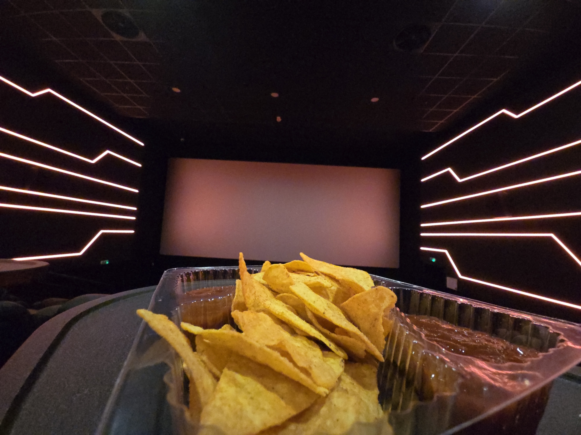 The image depicts a container of tortilla chips and two compartments of dipping sauce (likely salsa) on a seat in a movie theater. In the background, the theater screen is visible, illuminated and ready for a film. The walls are decorated with geometric neon light fixtures. The location is likely a cinema in Varna, Bulgaria, though no distinct features prove this. No people are visible. The scene suggests someone is at the movies with snacks.