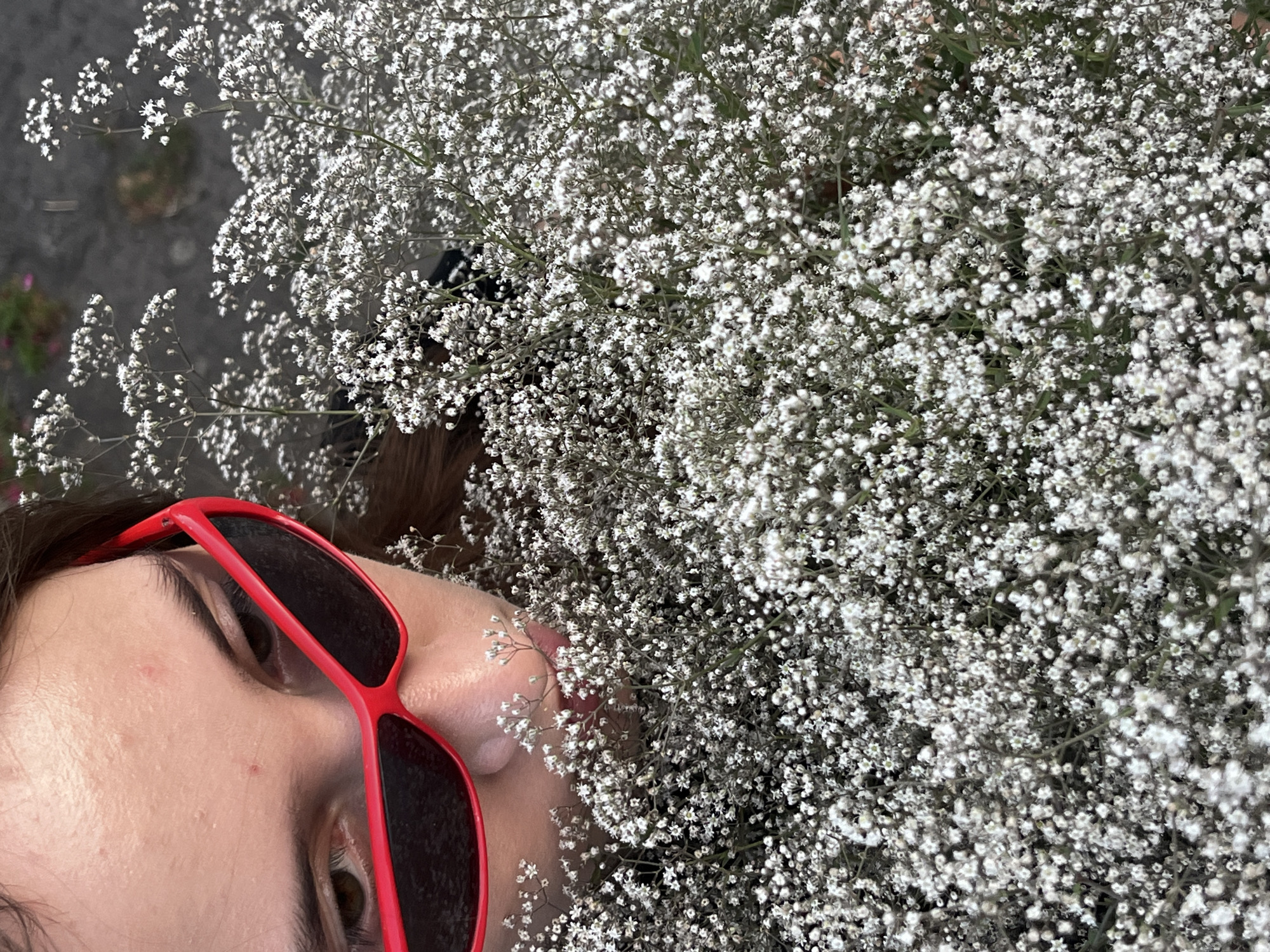 A close-up, eye-level, medium shot captures a person's face partially obscured by a large bouquet of small white flowers. The person wears bright red, angular sunglasses, and their brown eyes are visible through the lenses. Their skin is fair and appears to have some minor blemishes. The bouquet is dense and fills the majority of the frame, with the delicate white flowers and thin green stems creating a soft, textured backdrop. The background is softly blurred, showing hints of darker colors and indistinct shapes, suggesting an outdoor or urban environment. The lighting is natural, with soft shadows indicating an overcast or diffused light source.