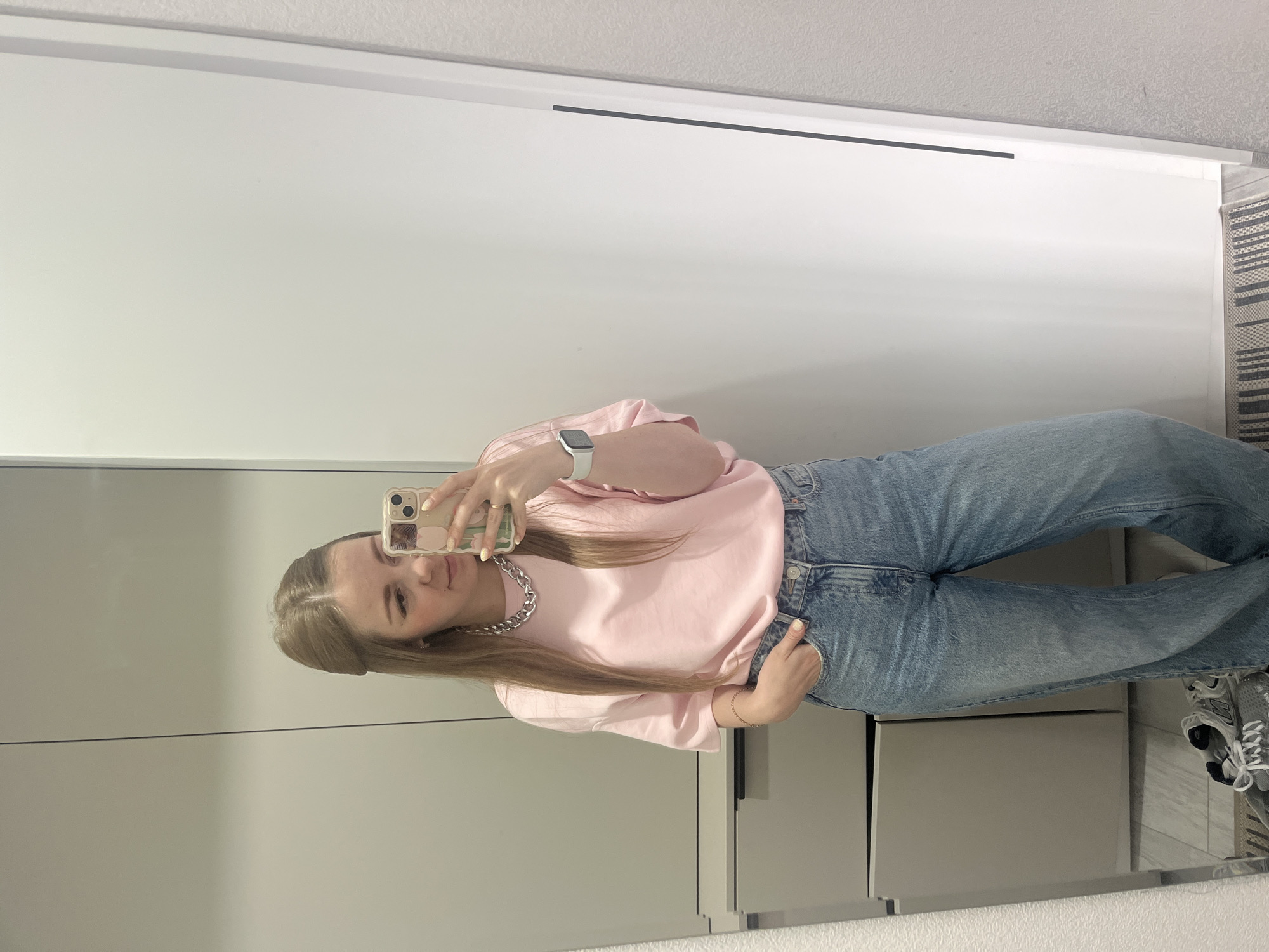 A blonde woman, approximately in her twenties, is centered in a mirror selfie, holding a light-colored smartphone with a floral-patterned case in her right hand. Her head is slightly tilted to her left, and she has visible winged eyeliner. Her long, light brown hair is parted in the middle, with the front sections pulled back. She is wearing an oversized, short-sleeved light pink t-shirt and light blue, loose-fitting denim jeans. A thick silver chain necklace is visible around her neck, and a light-colored smartwatch is on her left wrist. Her fingernails are painted yellow. Her left hand is partially in the front pocket of her jeans.
The reflection shows an interior space, likely a room or hallway. To the left of the woman, a light grey cabinet or closet unit with dark rectangular handles and drawers is visible. To her right, a plain white wall or door with a thin black vertical handle strip is seen. Further to the right, a light grey wall with a subtle textured pattern is present. The floor is light-colored, possibly tile or wood, with a dark and light striped rug positioned horizontally at the bottom of the frame. A pair of grey and white athletic sneakers is visible on the rug near the bottom left corner of the image. The scene is located in Unknown, Unknown.