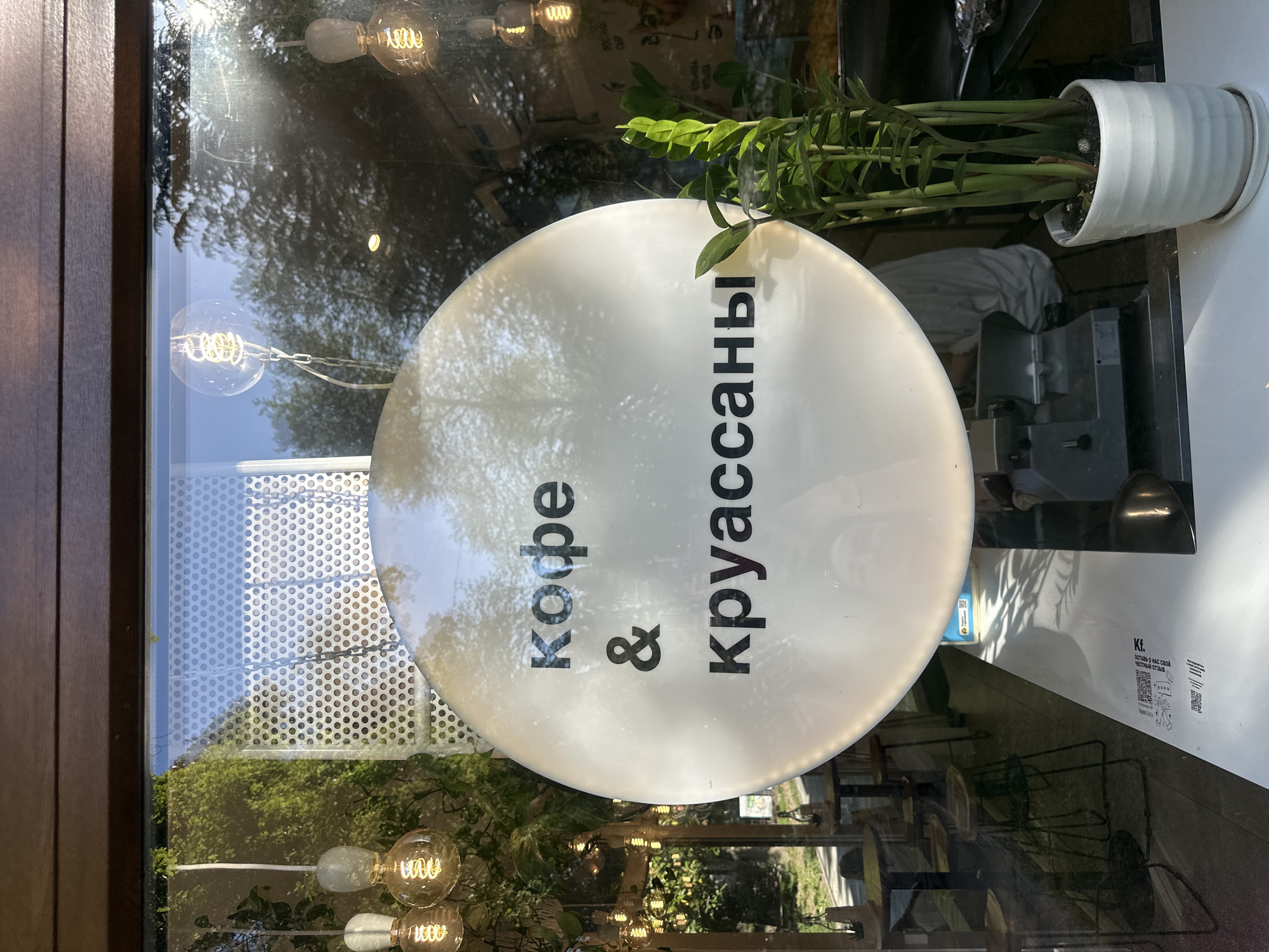 The image depicts a storefront window in Sochi, Russia, displaying a circular sign advertising "Кофе & круассаны" (Coffee & Croissants) in Cyrillic script. The sign is white and opaque, mounted on the window, partially obscuring the view of the interior and the outdoor environment.
Reflections on the window surface reveal the exterior environment: trees, foliage, and decorative lightbulbs suspended on strands. The foreground shows partial reflections of tables and chairs.
The interior, visible through the partially obscured glass, includes a potted plant with green foliage atop a white counter. A piece of equipment sits on the counter behind the plant. A white surface with printed information and QR codes is also visible inside the establishment.
The window's frame is dark brown.