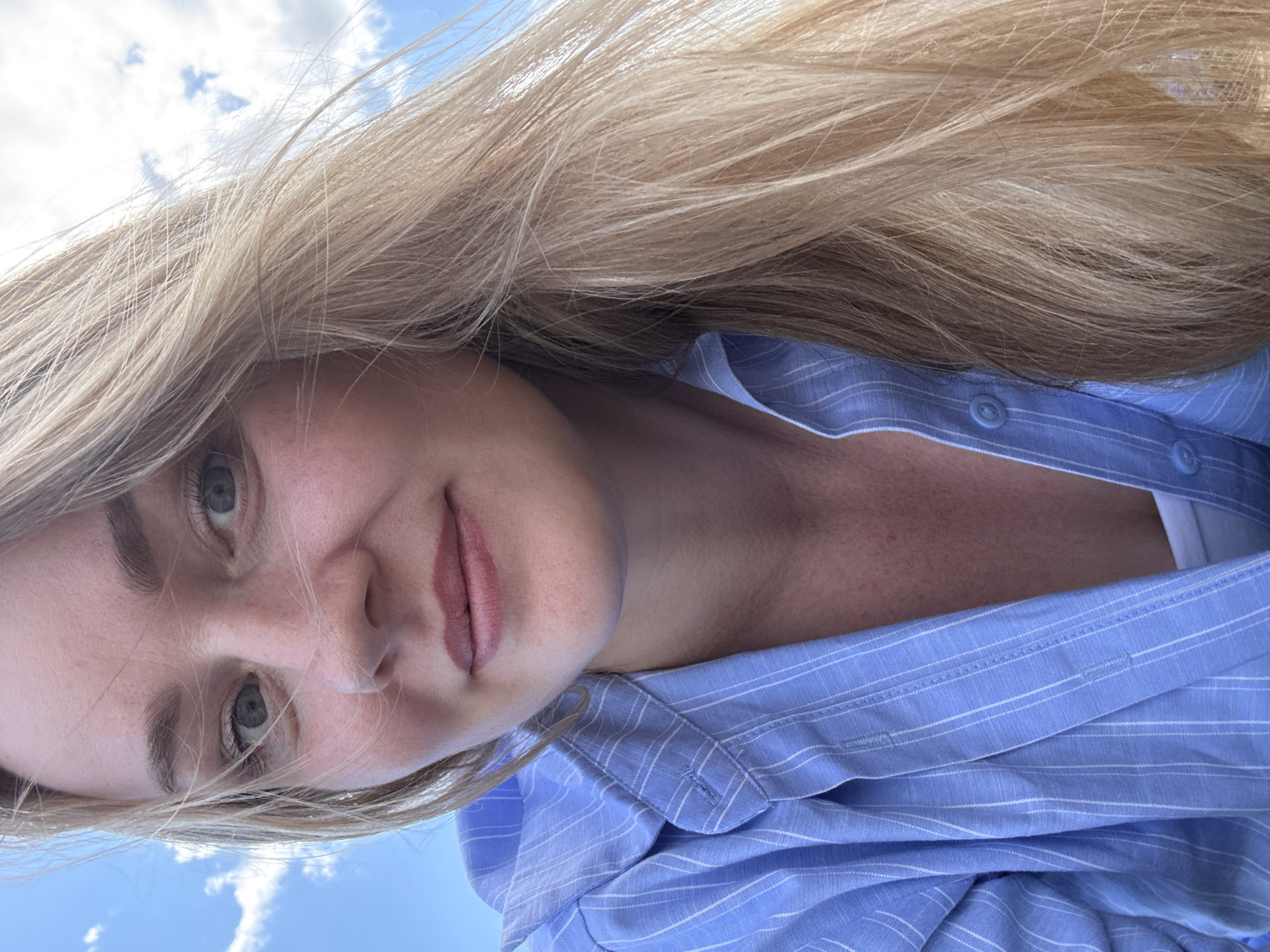 The media file is a close-up, upside-down selfie of a woman with blonde hair and fair skin. She is wearing a light blue, pinstriped button-up shirt, with the collar and some buttons visible. Her eyes are a light green, and she has light brown eyebrows and rosy lips. A strand of her blonde hair is falling across her face. The background is a bright blue sky with scattered white clouds, suggesting it is daytime and the weather is clear. The image appears to have been taken outdoors. No text is visible in the media file. The location context provided is Chaikovsky, Russia.