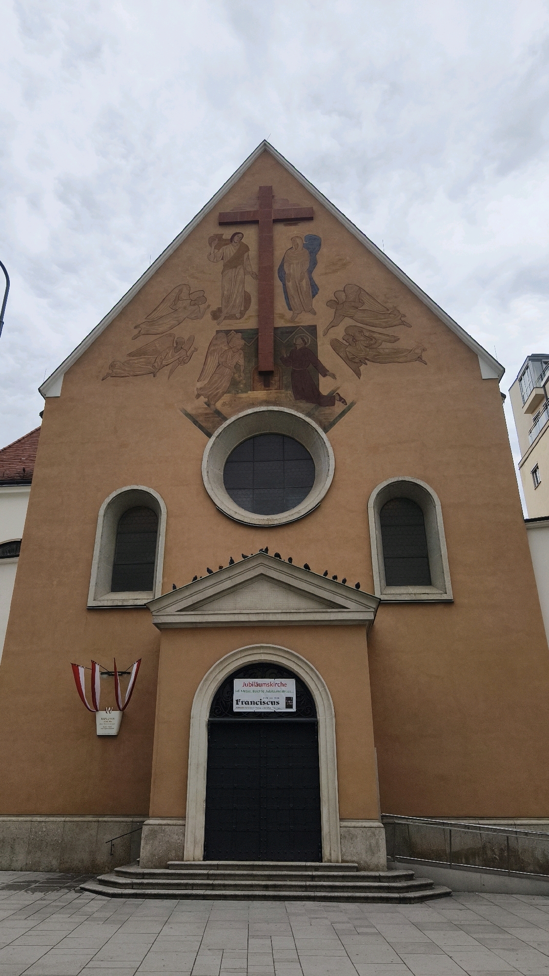 The image shows the facade of a church in Vienna, Austria. The church has a salmon-colored exterior with a large brown cross mounted on its triangular pediment, above a mural depicting figures and angels. Below the mural are three windows, a large circular one in the center and two arched ones on either side. A row of pigeons is perched on the ledge above the entrance. The entrance is an arched doorway with a large black door, flanked by two arched windows. A sign above the door reads "Jubiläumskirche" and "Franciscus." To the left of the entrance, a plaque with Austrian flags is mounted on the wall. The ground in front of the church is paved with light-colored tiles, and there are steps leading up to the entrance. The sky is overcast.