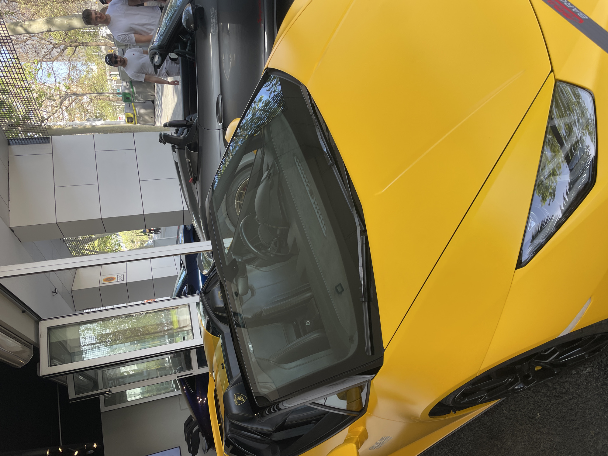 The image shows a yellow Lamborghini parked in front of a modern building in Barcelona, Spain. Two men are standing nearby a grey convertible car. The environment appears to be a showroom or a dealership. The architecture includes large glass windows. The cars are the focal point, with the yellow Lamborghini prominently displayed.