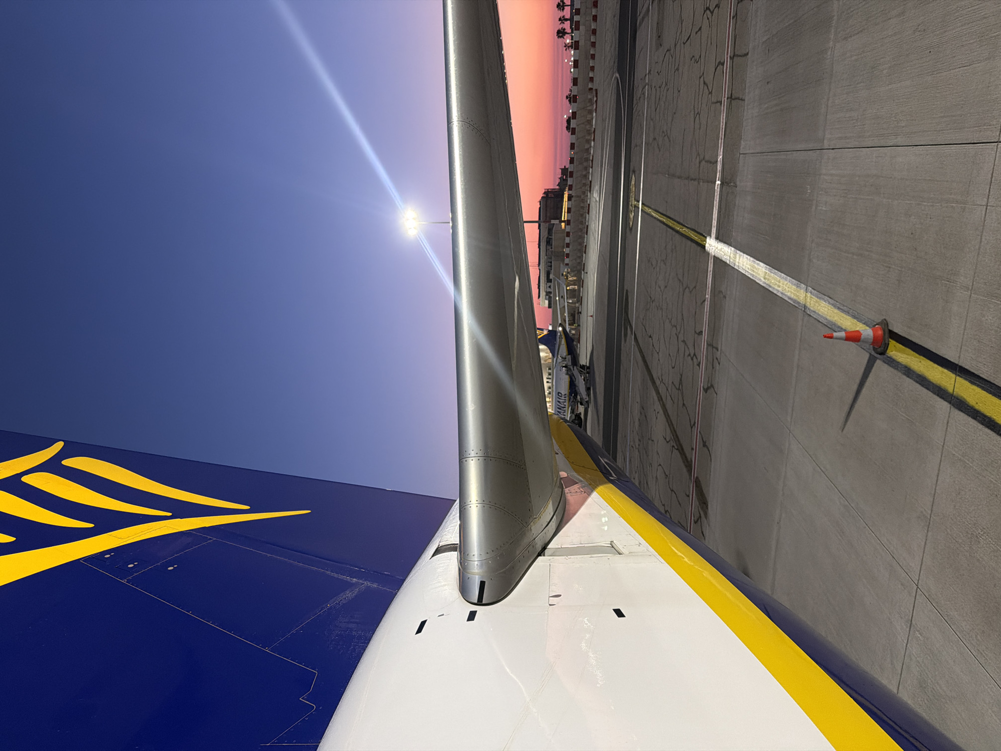 The image shows part of a Ryanair aircraft at Malta International Airport in Luqa, Malta. The tail of the aircraft is visible with the Ryanair logo in yellow on a blue background. The white wing with a yellow stripe is in the foreground, extending towards a grey concrete tarmac. A street lamp is visible above the plane, and a cone and a yellow line are seen in the background. There is a sunset with orange and pink hues in the background. Buildings and palm trees can be seen in the distance.
