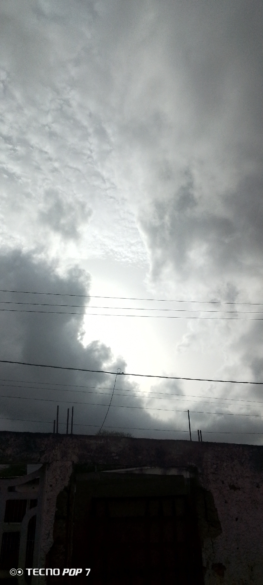 A low-angle, vertical outdoor shot captures a gloomy sky with heavy, dark clouds. The sky takes up most of the frame, with lighter clouds and a hint of sun breaking through in the upper middle. Below the sky, silhouetted against it, are several black power lines that stretch horizontally across the image. At the very bottom, the top of a low, weathered building is visible, with some broken masonry. To the left of the building, a portion of a white gate or fence can be seen. The bottom left corner of the image has a watermark that reads "TECNO POP 7". The scene appears to be in Altagracia, Venezuela.