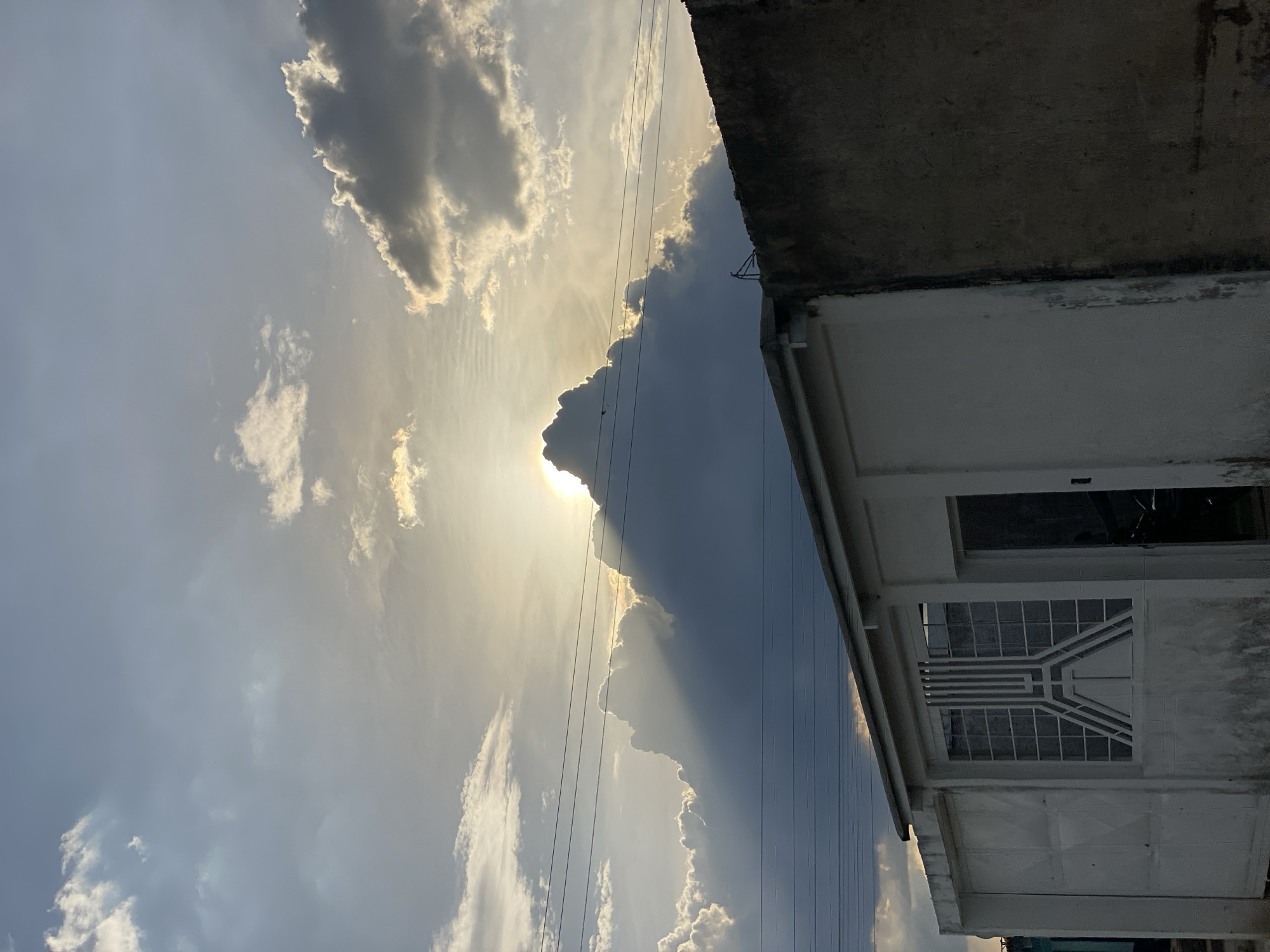 The image, taken in Tinaquillo, Venezuela, shows a low-angle view towards the sky, partially obscured by a building on the right. The sky contains a mix of light and dark clouds with the sun shining behind some of the clouds, creating rays of light. Power lines run across the view. The building is white with a window that has a grid-like pattern. The rooftop of the building is grey.