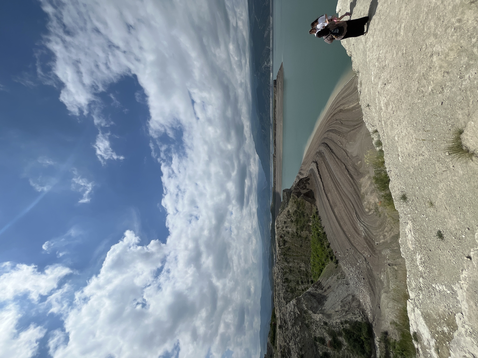 Two individuals are positioned on the edge of a steep cliff in Chirkey, Russia. The cliff overlooks a body of water and a landscape featuring layered geological formations. The sky is partially covered with white clouds.