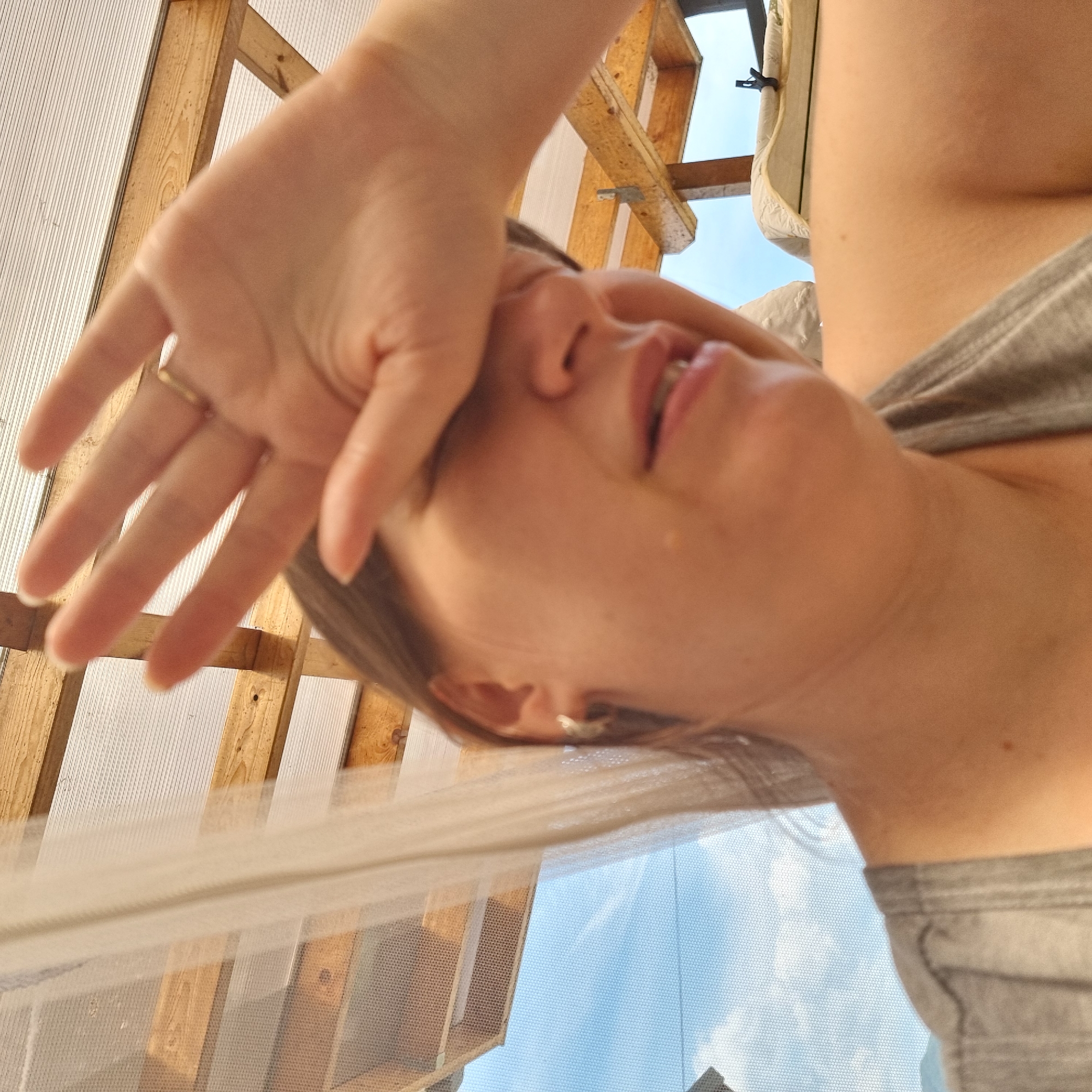 In a close-up, outdoor shot, a person is seen from the chest up, shielding their eyes with their right hand. The person is wearing a gray tank top and a small hoop earring in their visible left ear. Their facial expression is obscured, but their mouth is slightly open. Behind them, a translucent white fabric hangs, and a wooden structure with a corrugated plastic roof is visible. The background shows a blue sky with scattered clouds. The image appears to be taken on a sunny day. The location is not specified as Dunnae-myeon, South Korea.