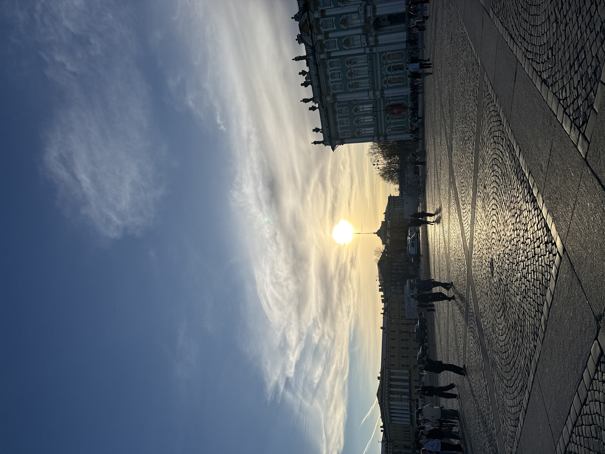 The image captures a sunny day at the Palace Square in Saint Petersburg, Russia. The Winter Palace is visible on the upper right side. A bright sun appears low in the sky, obscured by thin clouds. People are walking across the cobblestone square. The buildings line the lower portion of the image. The sky is blue with streaks of clouds.