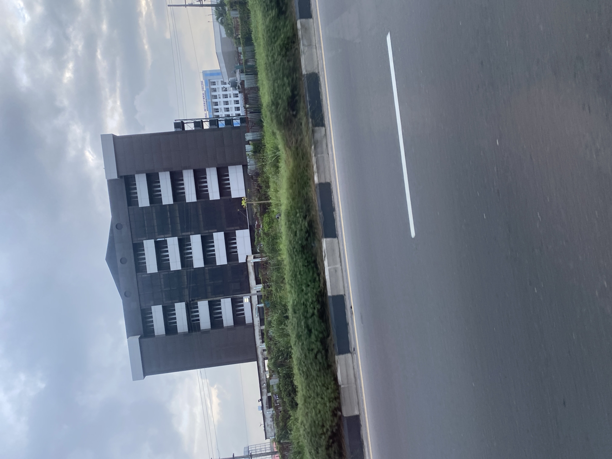 A medium shot captures a tilted view of a multi-story building with a dark facade and white window frames, set against a cloudy sky. In the background, a smaller, lighter-colored building with the text "GLOBAL INTERNATIONAL COLLEGE" is visible. The foreground features a road with a white line, a median with black and gray dividers, and lush green vegetation bordering the road. The scene appears to be a roadside view of urban architecture and infrastructure under an overcast sky.