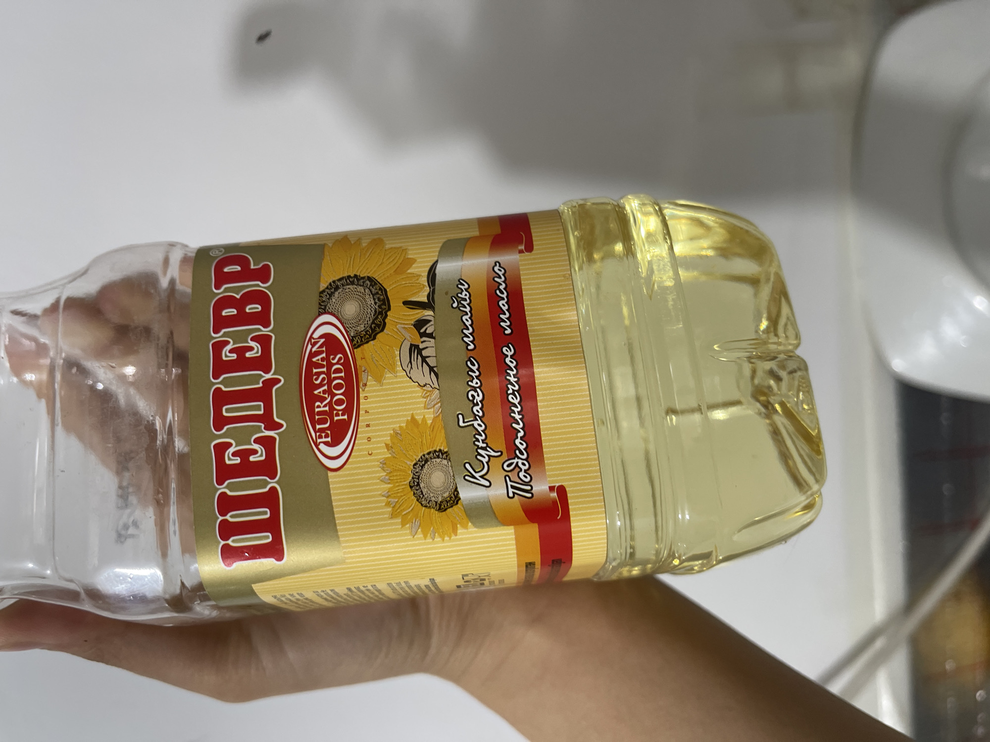 A hand holds a clear plastic bottle of sunflower oil. The label on the bottle is predominantly gold with red and yellow accents. The Russian word "ШЕДЕВР" (Shedevr) is prominently displayed in red lettering with a white outline. Below this, "EURASIAN FOODS" is written in white within a red oval. The label also features images of sunflowers and text in a cursive font, including "Кунбагыс майы" and "Подсолнечное масло". The oil inside the bottle is a pale yellow color. The background is a plain white wall with some light and shadow. The scene appears to be indoors, possibly in a kitchen or pantry, in Bishkek City, Kyrgyzstan.