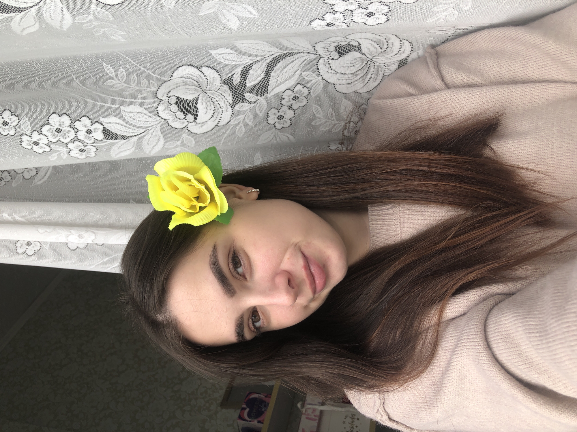 This is a portrait-style image of a young woman, her head tilted to her right, looking directly at the viewer with a gentle, soft smile. She has long, straight, dark brown hair that falls over her shoulders and part of her light-colored, possibly beige or light pink, knitted sweater. A vibrant yellow artificial flower, resembling a rose, with a green leaf, is tucked behind her right ear. She wears a small, silver-toned earring in her visible ear.
The primary background element is a white lace curtain, richly patterned with floral motifs, including roses and leaves. Below the curtain, on the left side of the frame, a dark, subtly patterned surface is visible. In the blurred background to the right of the woman, various household items are indistinctly visible on shelves, reinforcing a home environment. On a pink object within this blurred background, the Cyrillic word "ЛЮБЛЮ" (meaning "I love") and a heart symbol are partially discernible. The woman's expression conveys a calm and friendly demeanor. The scene takes place in what is identified as Noviy, Russia.