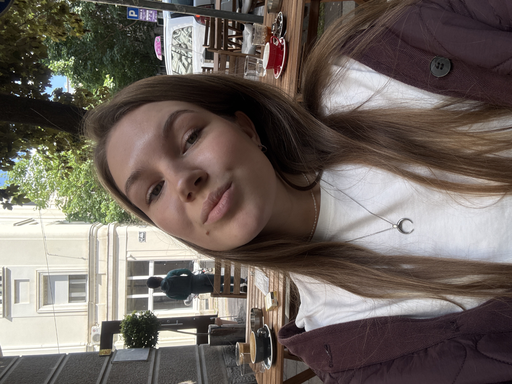 A young woman is seated at an outdoor cafe in Belgrade, Serbia. She is wearing a white t-shirt, a brown jacket, and a silver necklace with a crescent moon pendant. She has light brown hair and appears to be taking a selfie. On the wooden table in front of her, there are cups and saucers, indicating she is having coffee or another beverage. In the background, there is a building with white trim, a person in a green jacket walking by, trees, and a vehicle with a parking sign nearby, suggesting a typical urban street scene in Belgrade.
