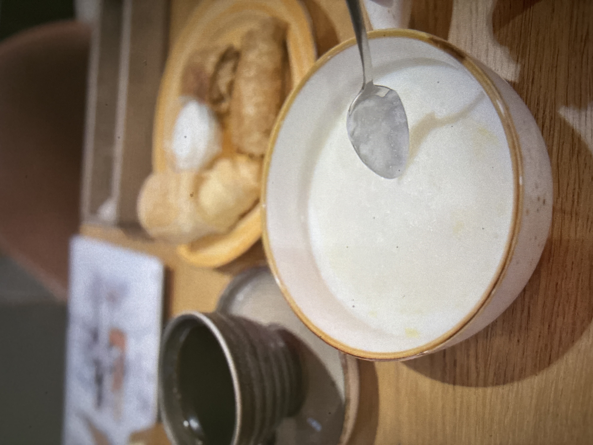 An overhead, close-up shot shows a breakfast scene on a wooden table in Novosibirsk, Russia. In the foreground, a light-colored bowl with an orange rim contains a thick, white porridge. A silver spoon rests in the porridge, its bowl angled upwards. To the left and slightly behind the porridge bowl, a dark-colored mug with a ribbed texture sits on a matching saucer. The mug is filled with a dark liquid, likely coffee. In the background, out of focus, a yellow plate holds two golden-brown pastries, one topped with a dollop of white cream. The image has a grainy texture.