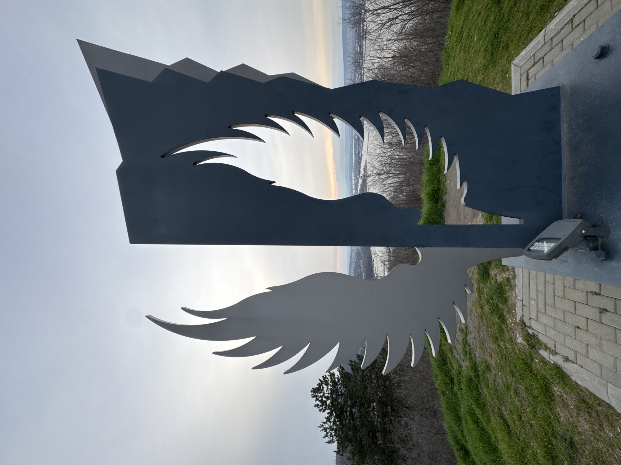 The image shows a metal sculpture in Varna, Bulgaria. The sculpture features a stylized mountain range at the top with cut-out shapes resembling eagle wings. A solid metal wing extends from the sculpture on the bottom half. In the background, there's a landscape view of the city and the Black Sea. The sculpture is located on a paved area with grass and trees nearby. A light fixture is attached to the sculpture.