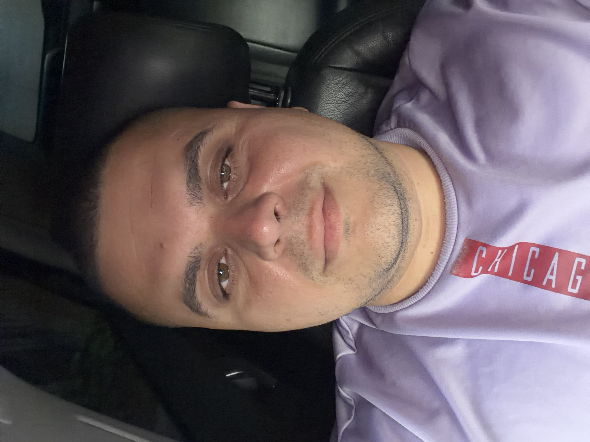 A man is depicted in a close-up, tilted self-portrait, likely taken within a car. He has short, dark hair and light stubble on his chin and cheeks. His eyes are brown, and he looks directly towards the camera with a neutral, calm expression. His skin tone is fair to medium, and a slight sheen suggests either natural skin texture or low-light reflection.
He is wearing a light purple, crew-neck sweatshirt or t-shirt. On the front of his shirt, partially visible on the right side of the image, is red text that reads "CHICAG", which appears to be part of the word "CHICAGO".
The background suggests an indoor setting, specifically a vehicle interior. A dark, textured headrest or car seat is visible behind and above his head, indicating he is seated. Other dark, indistinct elements of a car's interior, possibly parts of the dashboard or door, are visible around him. The lighting is soft and somewhat dim, possibly ambient light from the vehicle or evening hours.
The image captures a casual moment of the man in what appears to be a car in Shumen, Bulgaria.