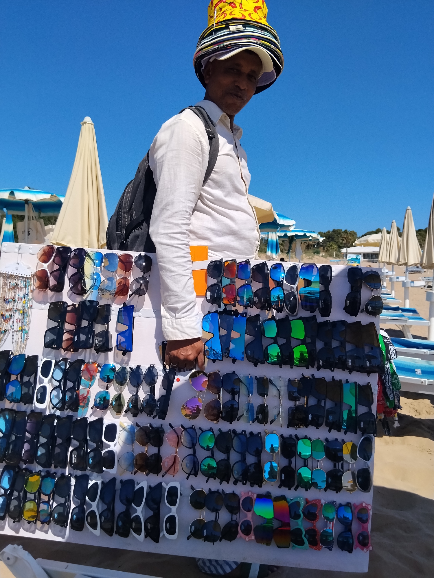 In Peschici, Italy, a man stands on a sandy beach, facing slightly left, and wearing a tall, multi-layered hat adorned with various colorful caps and a yellow, chili-patterned element on top. He is dressed in a light-colored, long-sleeved shirt and a dark backpack is slung over his shoulders. In front of him, a white display board is set up, showcasing a large assortment of sunglasses in various styles, colors, and lens tints. The sunglasses are neatly arranged in rows, attached to the board. In the background, beach umbrellas and lounge chairs are visible, suggesting a typical beach scene under a clear blue sky. The man appears to be selling the sunglasses to beachgoers.
