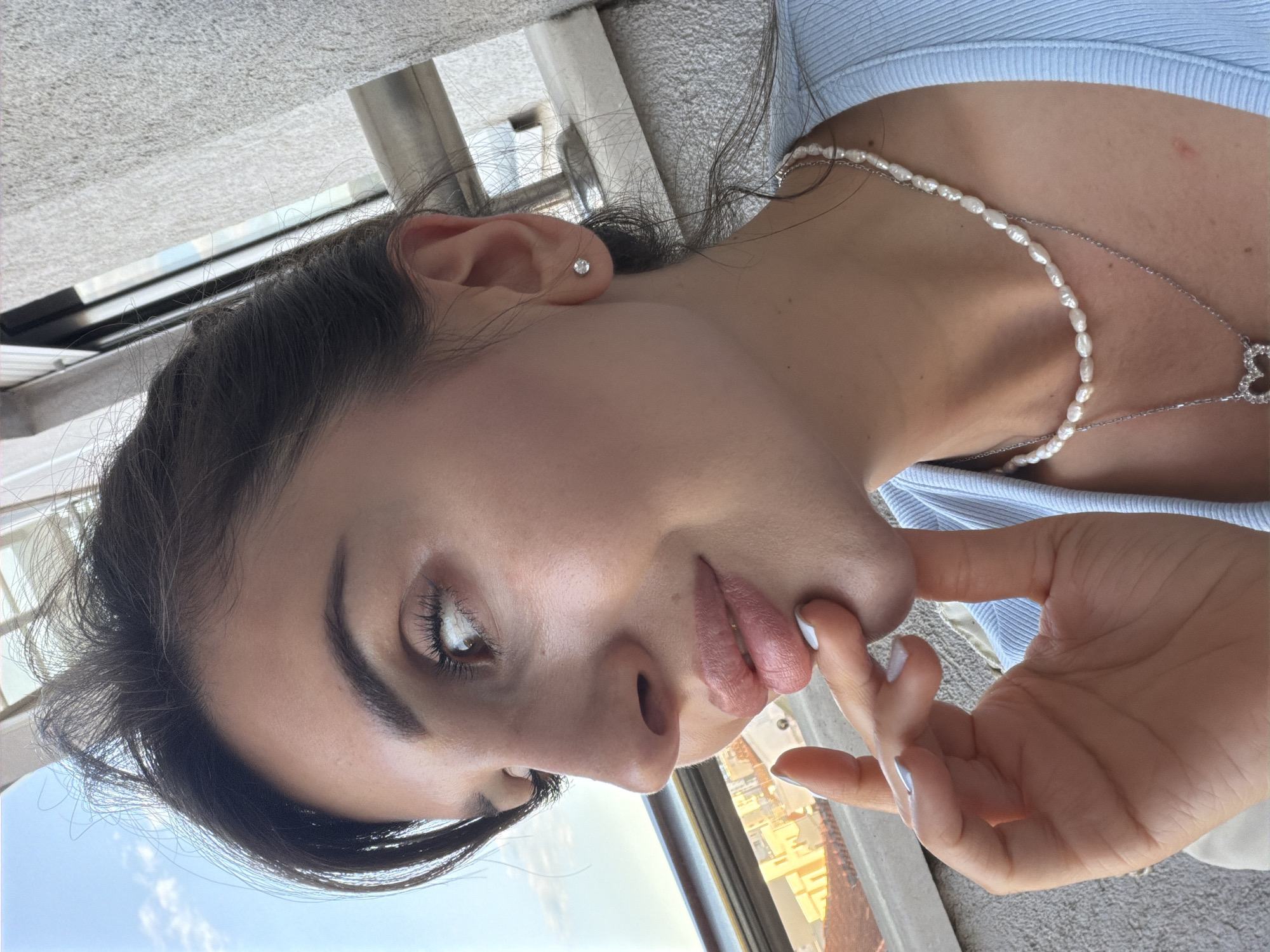 A medium shot depicts a woman's face and upper chest from a slightly elevated perspective. She has dark hair, fair skin, brown eyes, and is wearing a light blue top. A pearl necklace and a silver chain with a heart pendant adorn her neck. Her left hand is raised, with her fingers gently touching her lips. The background shows an exterior scene, likely taken from a high-rise building, revealing a section of the city with clear skies, buildings, and the horizon. This view suggests the photo was taken in a city setting, potentially in Budva, Montenegro, based on the architecture and coastal proximity seen in the distance.