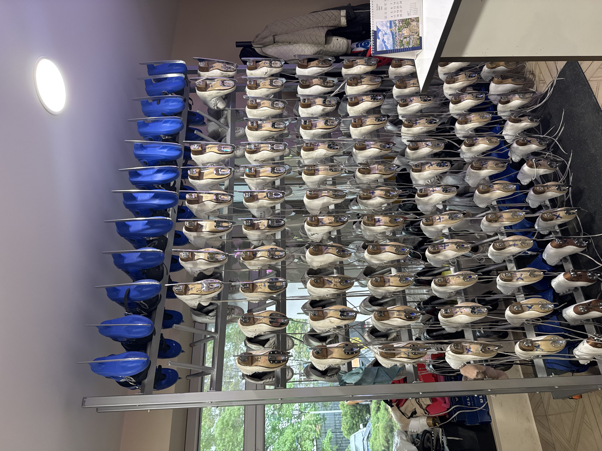 The image depicts a storage rack filled with figure skates. The rack is metal and has multiple levels. On one side, the skates are primarily blue, while on the other side, they are mostly white and tan. Some skates have identifying markings or labels on the blades. A calendar is visible on top of the rack and various other equipment. The location is Tbilisi, Georgia.