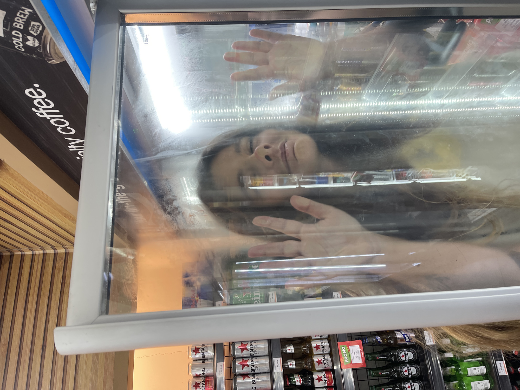 The media file is a photograph taken at a slightly low angle, featuring a person's face and hand pressed against a glass refrigerator door. The person appears to be a young adult with long, dark hair. They are looking upwards, with their eyes closed and a relaxed expression. Their left hand is outstretched, with fingers splayed against the glass. Behind the person, the refrigerator is filled with various beverages, including cans of Bintang beer and bottles of Heineken. The lighting inside the refrigerator is bright and cool, suggesting it is a well-lit commercial space.
On the left side of the image, a portion of a black sign with white text is visible, reading "COLD BREW", "ZERO SUGAR", "VEGAN", and "alty coffee.". The overall scene appears to be inside a store or convenience shop, likely in a warm climate given the presence of beer. The reflection on the glass door is prominent, showing a distorted view of the surroundings. The image seems to capture a candid moment of someone interacting with the refrigerator.