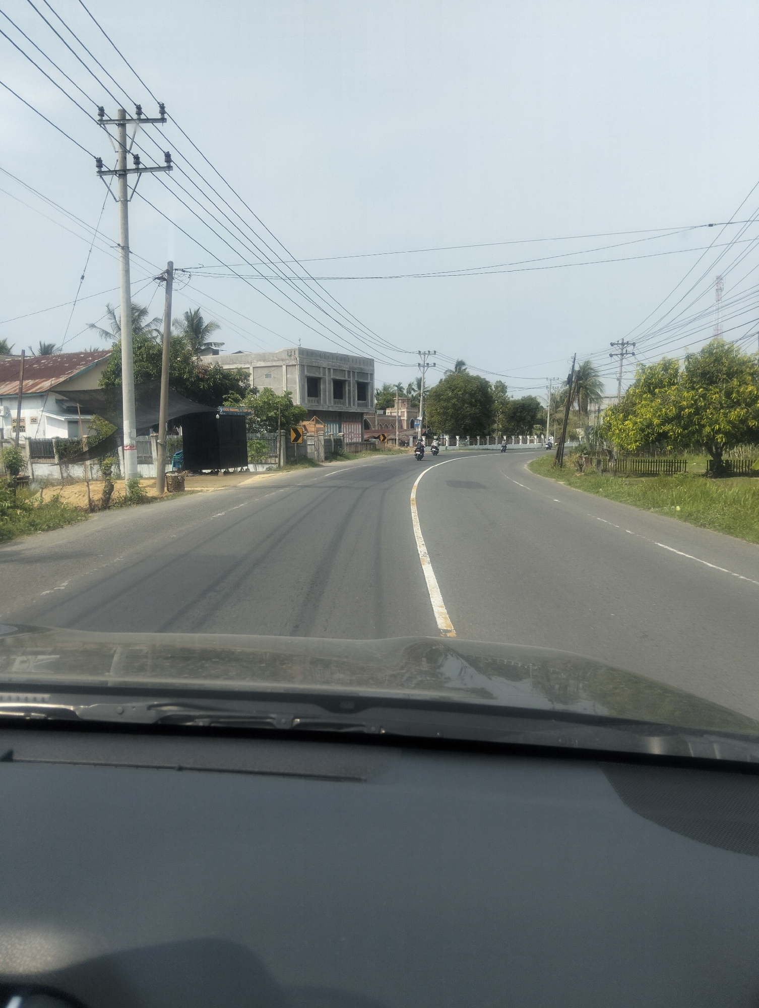 The image depicts a rural road in Cotabato, Indonesia, under an overcast sky. The road curves to the right, with a white line marking the center. On the left side of the road, there are houses and utility poles with numerous wires. A black structure, possibly a shop, is situated near the road. Further down the road, a partly constructed building is visible. On the right side of the road, there are trees and a fence. Several motorcycles are seen on the road, suggesting ongoing traffic. The foreground shows the interior of a vehicle, with the dashboard and windshield wipers visible.