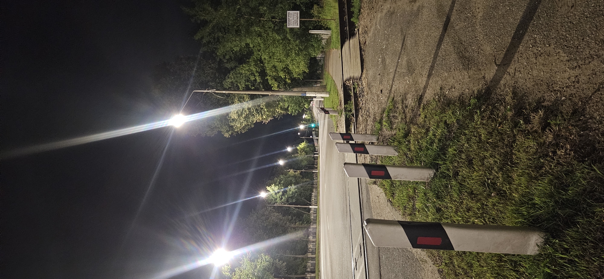The image captures a nighttime street scene with several streetlights casting bright beams across a paved road and grassy verge. In the foreground, a row of bollards with alternating black and white segments and red reflective markers lines the side of the road. Beyond these, a set of railway tracks crosses the road, with a wooden crossing. The road stretches into the distance, bordered by trees on both sides. In the background, more streetlights illuminate the scene, and a traffic light can be seen in the distance, appearing green. A signpost with text in Cyrillic is visible on the right side of the image. The overall impression is a well-lit urban or suburban road at night. The signpost reads: "ПЕШЕХОДНЫЙ ПЕРЕХОД. КАРТЫ ИЛИ ПОЕЗДА. ЗАПРЕЩЕНО!". This translates to: "PEDESTRIAN CROSSING. CARDS OR TRAINS. PROHIBITED!". The presence of the railway crossing and the sign suggests this is a railway crossing for pedestrians, with the prohibition likely referring to crossing the tracks when a train is approaching or present. This location is likely near the railway crossing on Moskovsky Prospekt in Vitebsk, Belarus, given the overall visual characteristics and the Cyrillic signage.
