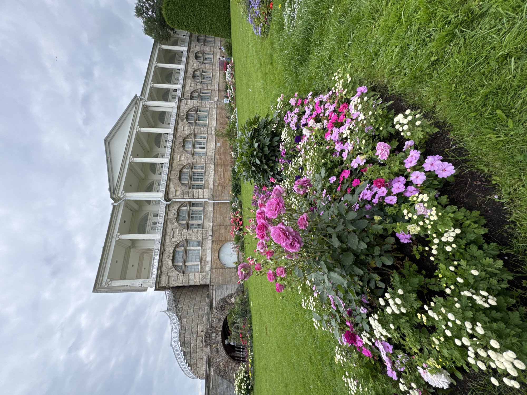 This image depicts the Cameron Gallery, a pavilion in Catherine Park in Pushkin, Russia. The pavilion is a neoclassical structure with a long colonnade of white columns and arched windows. In the foreground, a vibrant flowerbed bursts with pink roses, purple impatiens, and white chrysanthemums, set against a lush green lawn. To the left, a curved stone staircase with white railings leads up to the pavilion. The sky is overcast with grey clouds. Several people are visible in the distance, strolling along the colonnade and the park grounds. A tall, conical evergreen tree stands to the right of the pavilion.